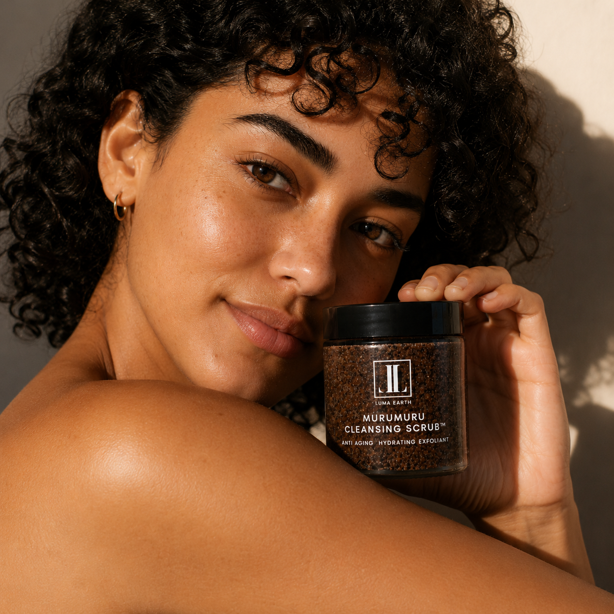 A woman with curly dark hair holding a jar of Luma Earth Murumuru Cleansing Scrub close to her face, looking at the camera with a slight smile, in warm lighting.