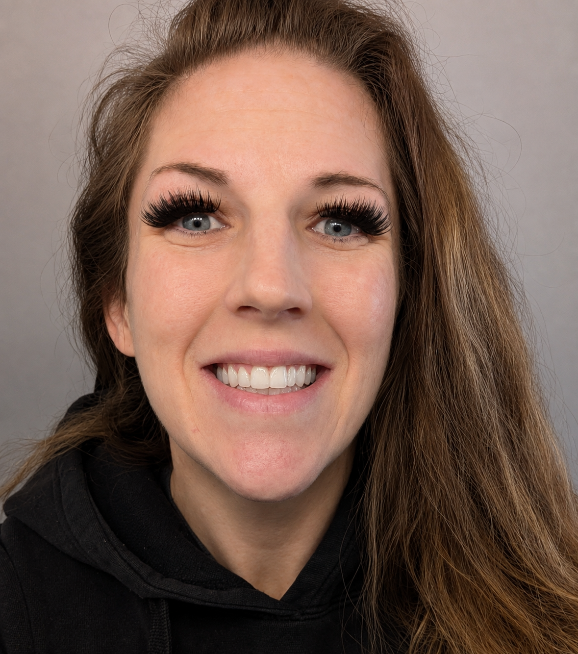 A smiling woman with long brown hair, wearing thick false eyelashes and a black hoodie, against a gray background.