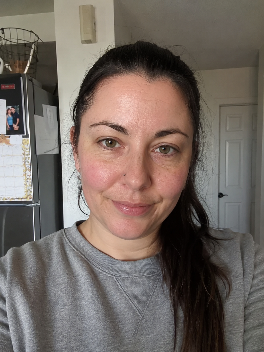 A woman with long dark hair tied back, wearing a gray sweatshirt, standing inside a house in front of a kitchen counter and a closed white door.