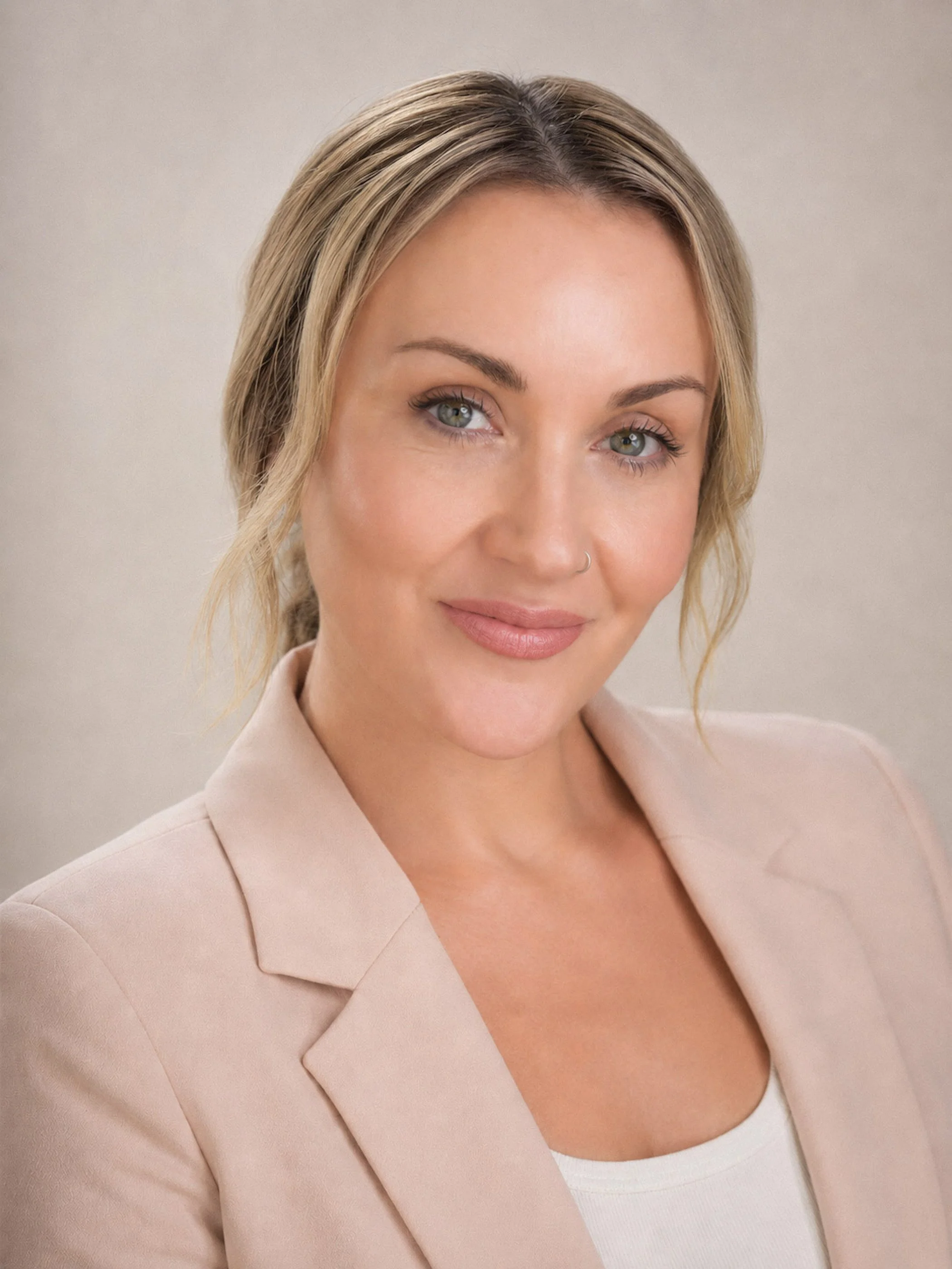 Close-up of a woman with blonde hair, light skin, green eyes, wearing a beige blazer and white top, smiling softly against a plain background.