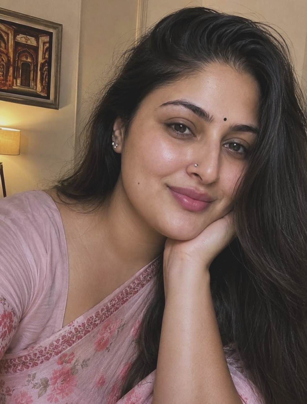 A woman with long dark hair and a nose piercing resting her face on her hand, wearing a pink saree with floral embroidery, indoors with a framed artwork and a lamp in the background.