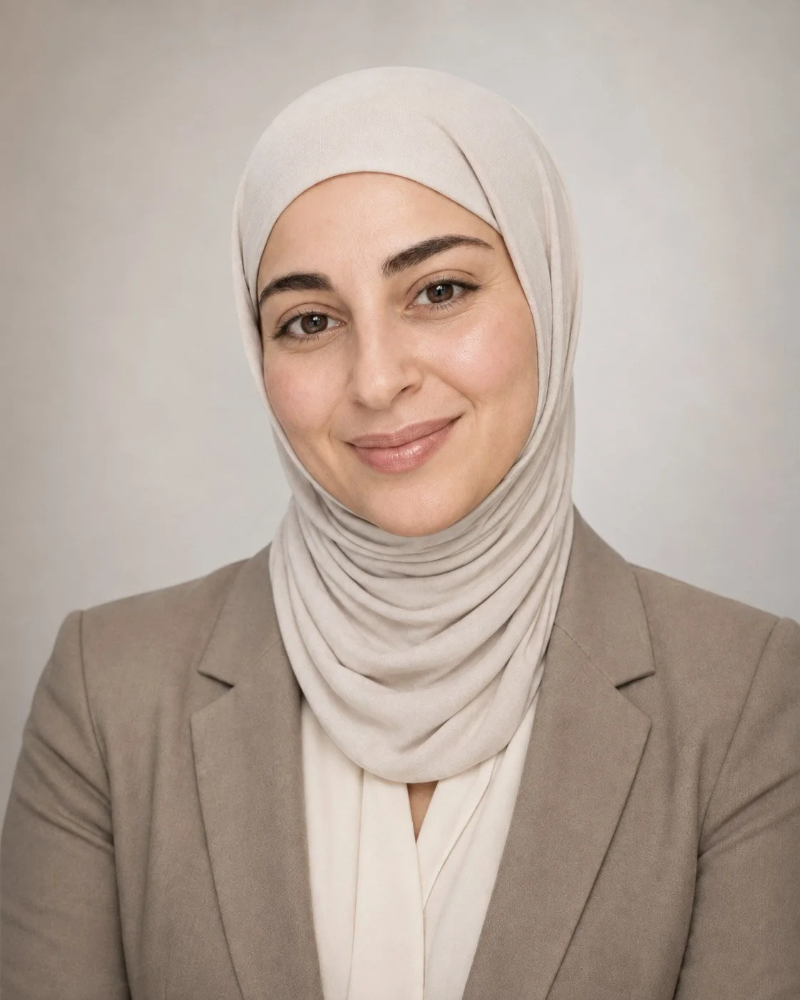 A woman wearing a beige hijab and a beige blazer, smiling at the camera against a plain background.
