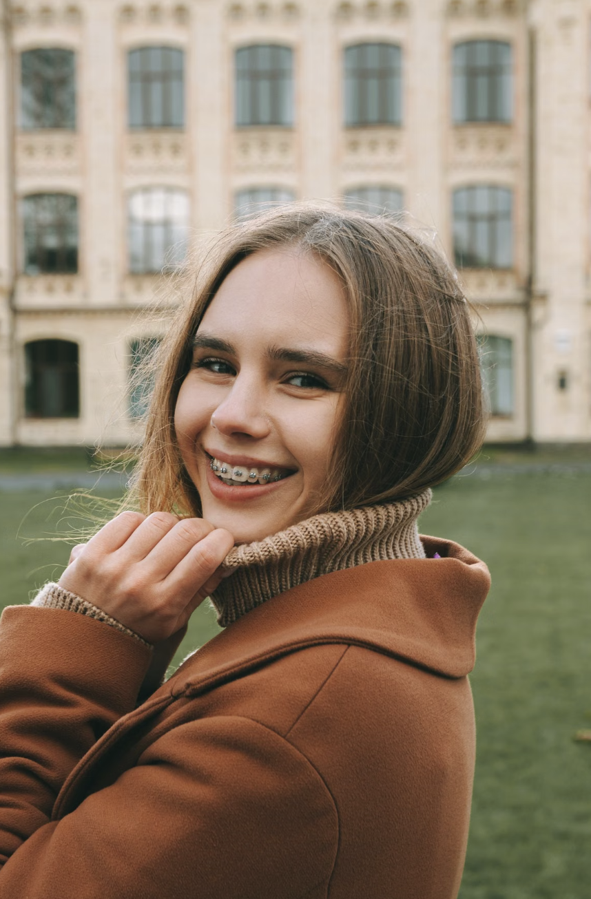 Young woman smiling outdoors, wearing a brown jacket and braces, with a historic building in the background.