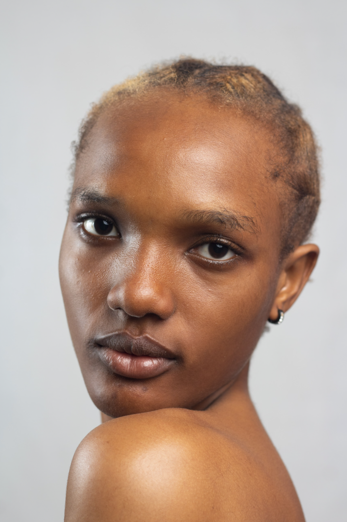 Close-up of a young woman with short, natural hair and medium brown skin, looking directly at the camera with a neutral expression, against a light gray background.