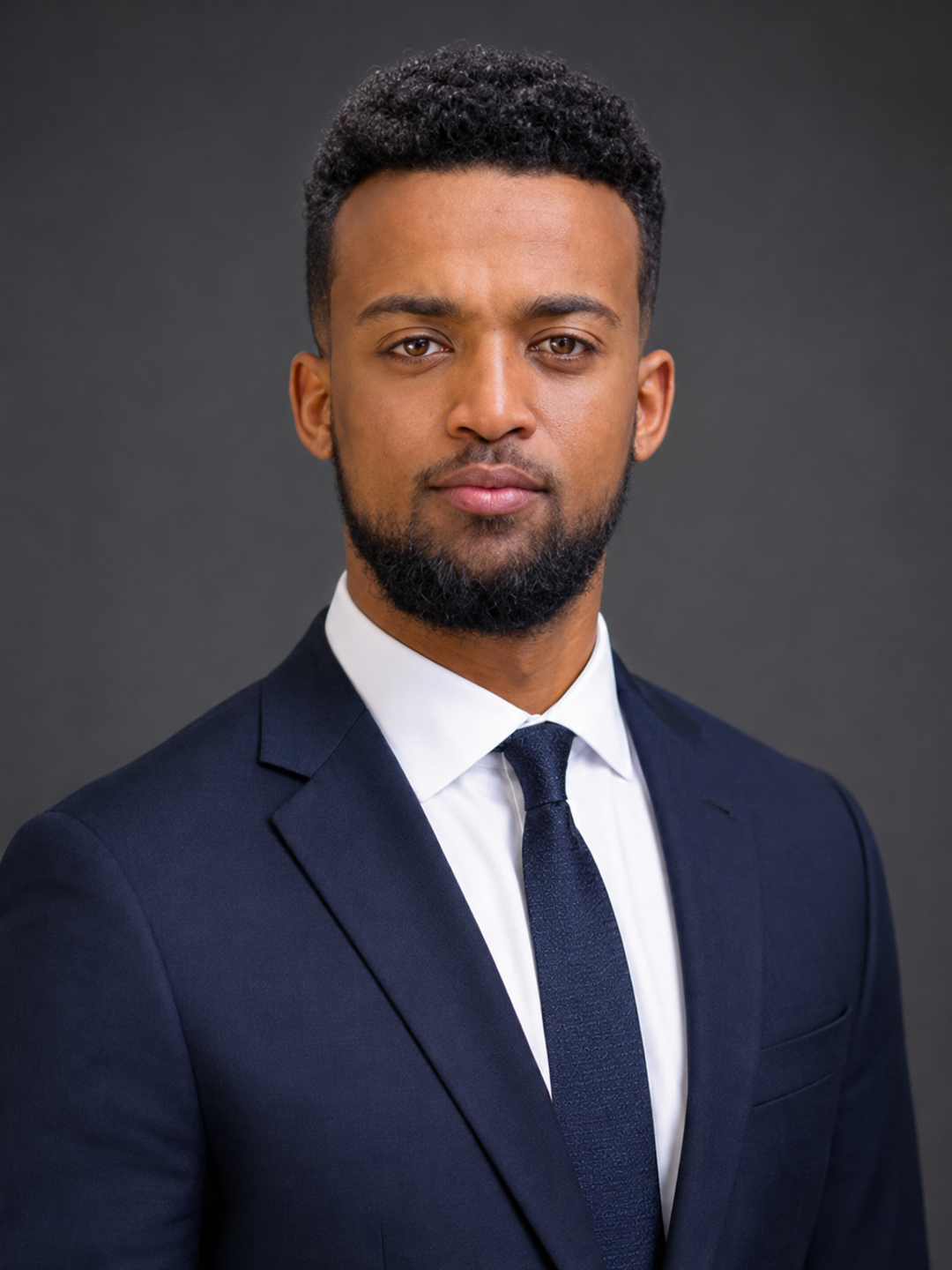 A young man with short curly hair, a beard, wearing a dark blue suit, white shirt, and dark tie, standing against a gray background.