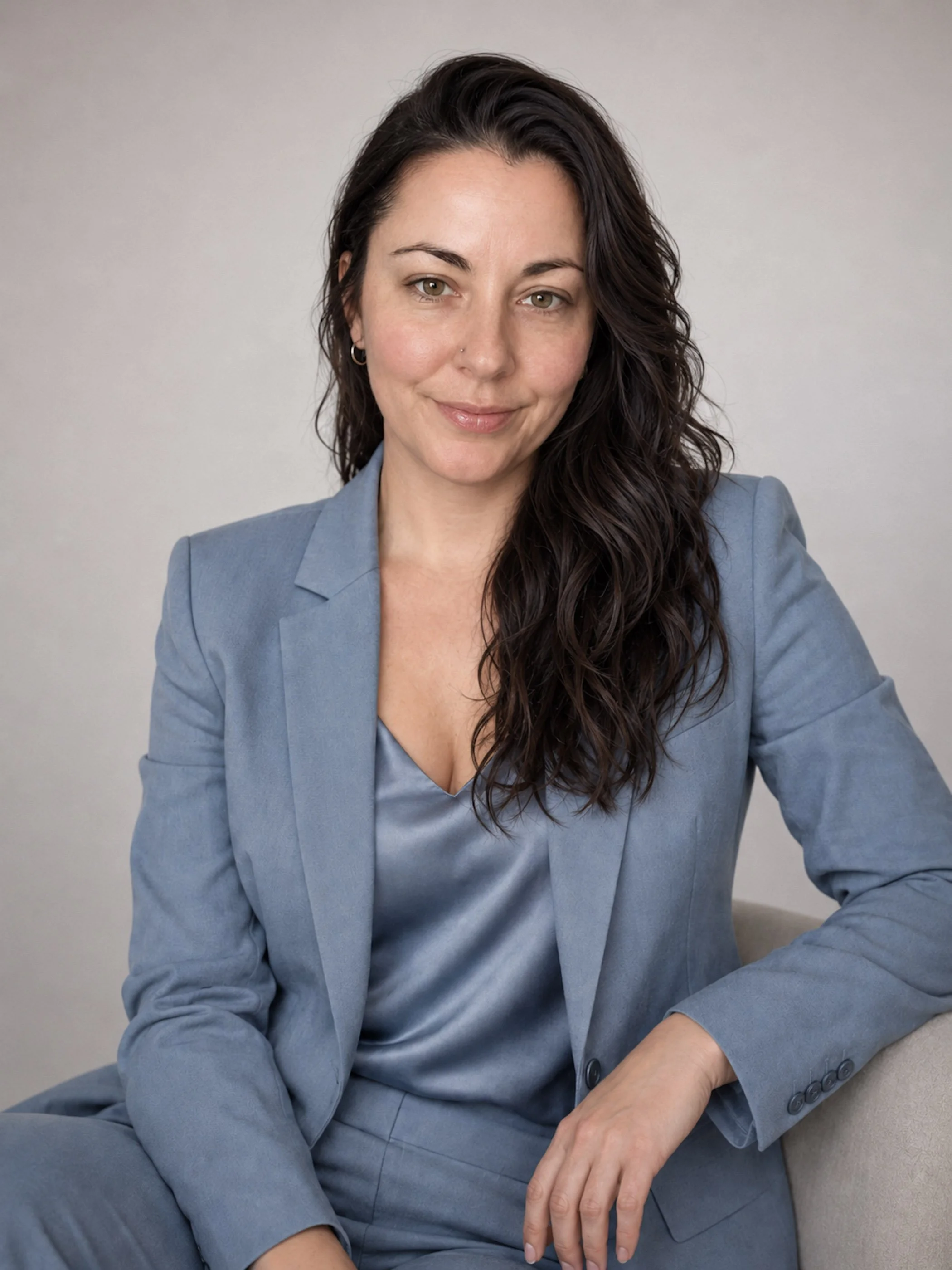 A woman with long dark wavy hair, wearing a light blue suit, sitting on a couch, smiling softly at the camera, with a plain background.
