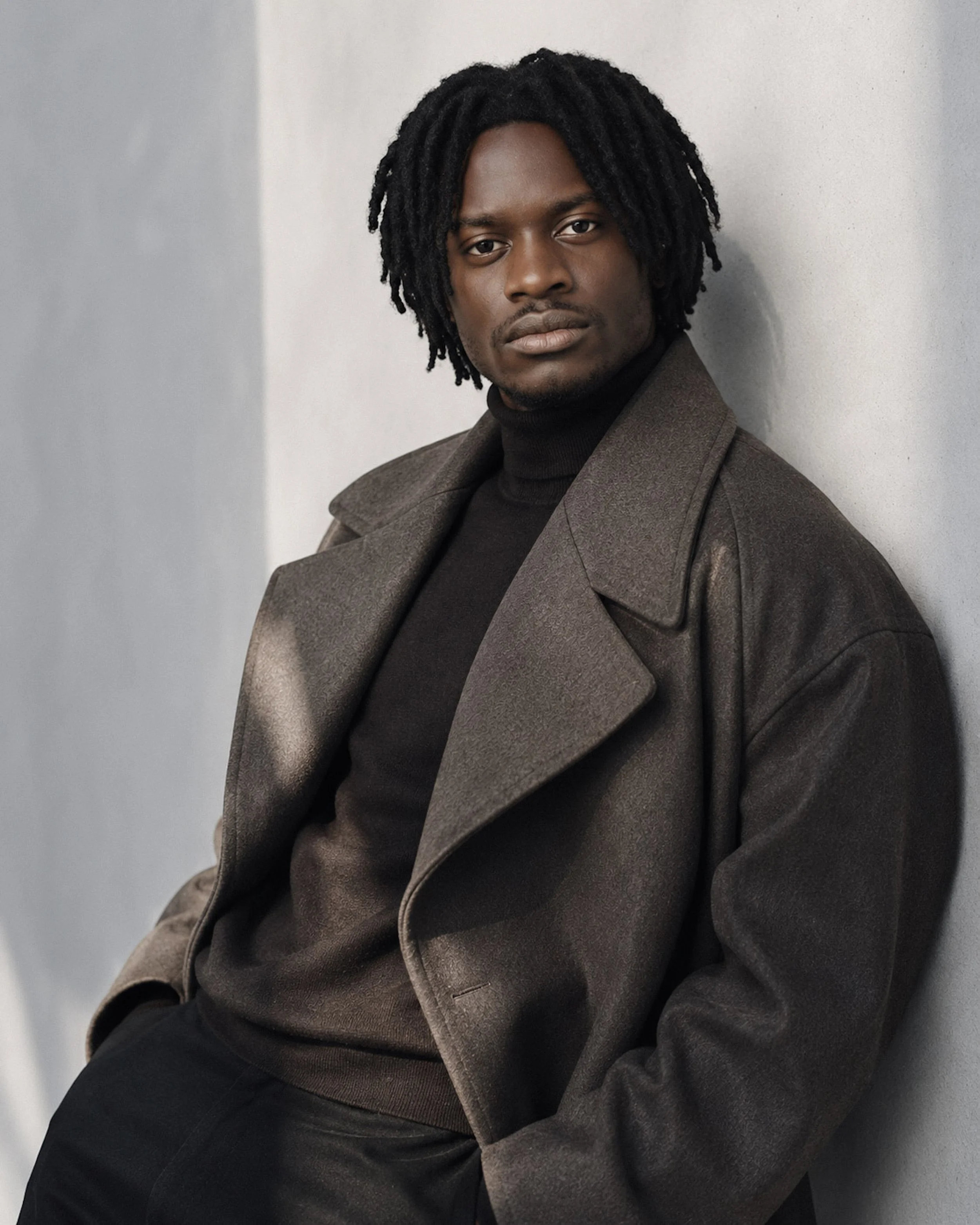 A man with dark skin and dreadlocks posing against a plain wall, wearing a brown coat and black turtleneck.