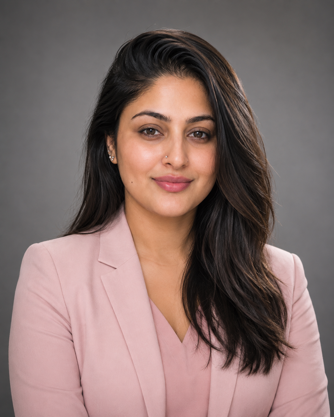 A woman with long dark hair and light skin, wearing a light pink blazer, smiling against a plain gray background.