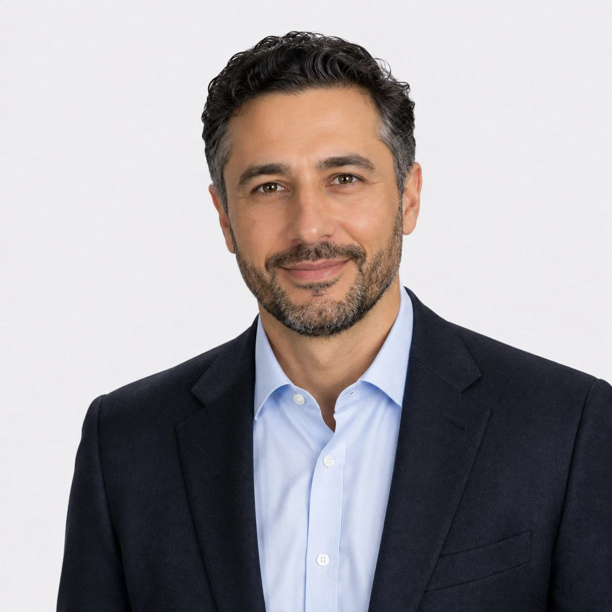 Professional headshot of a middle-aged man with dark, wavy hair and a beard, wearing a navy blazer and light blue dress shirt against a plain white background.