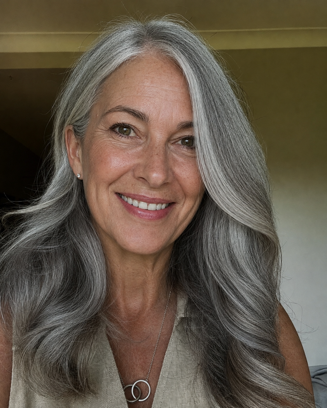 A smiling woman with long, wavy gray hair and light skin, wearing a beige sleeveless top, diamond stud earrings, and a silver necklace with a circular pendant, indoors with a greenish wall background.