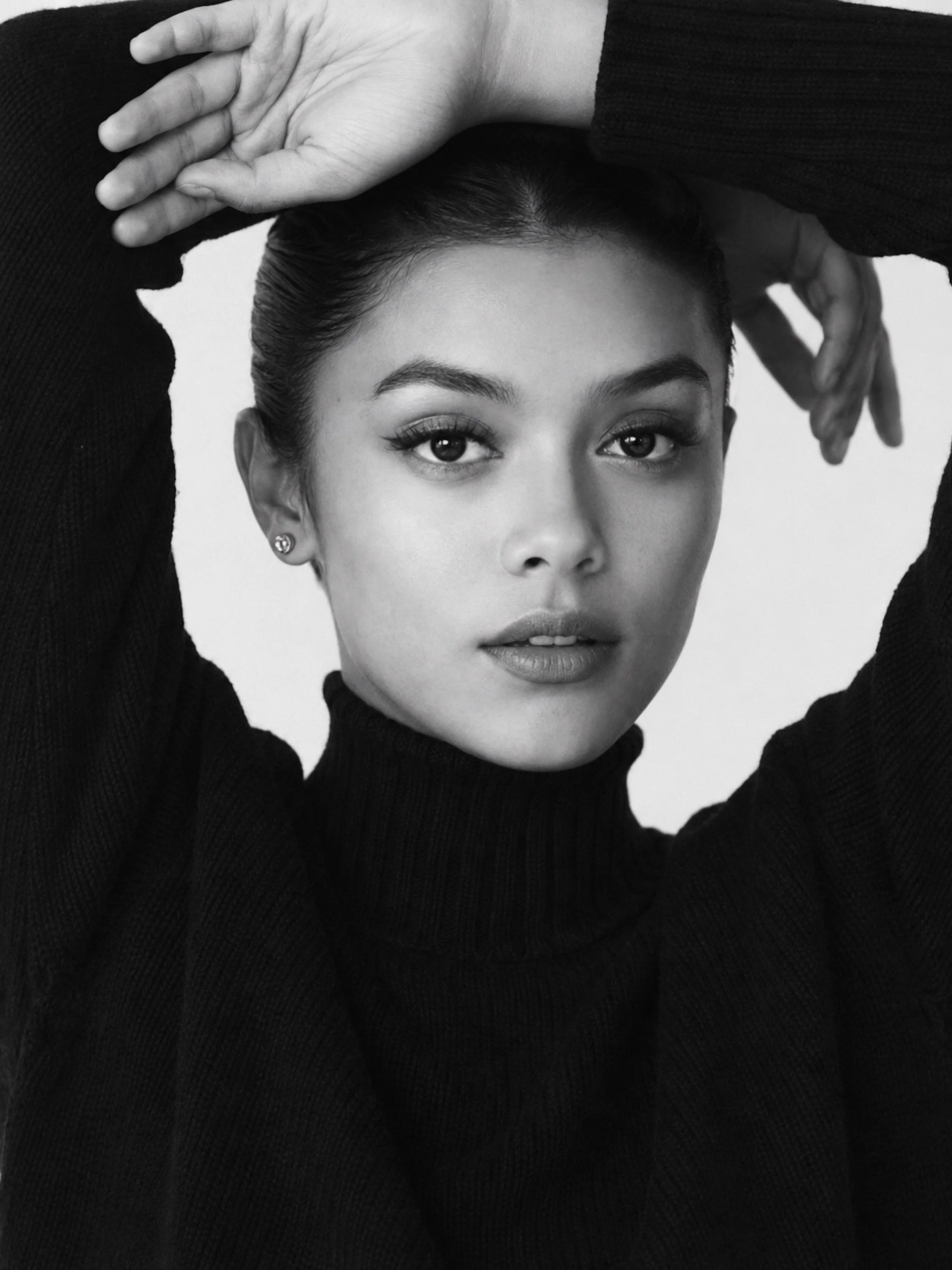 Black-and-white editorial portrait of a woman in a black turtleneck posing with her arms raised.