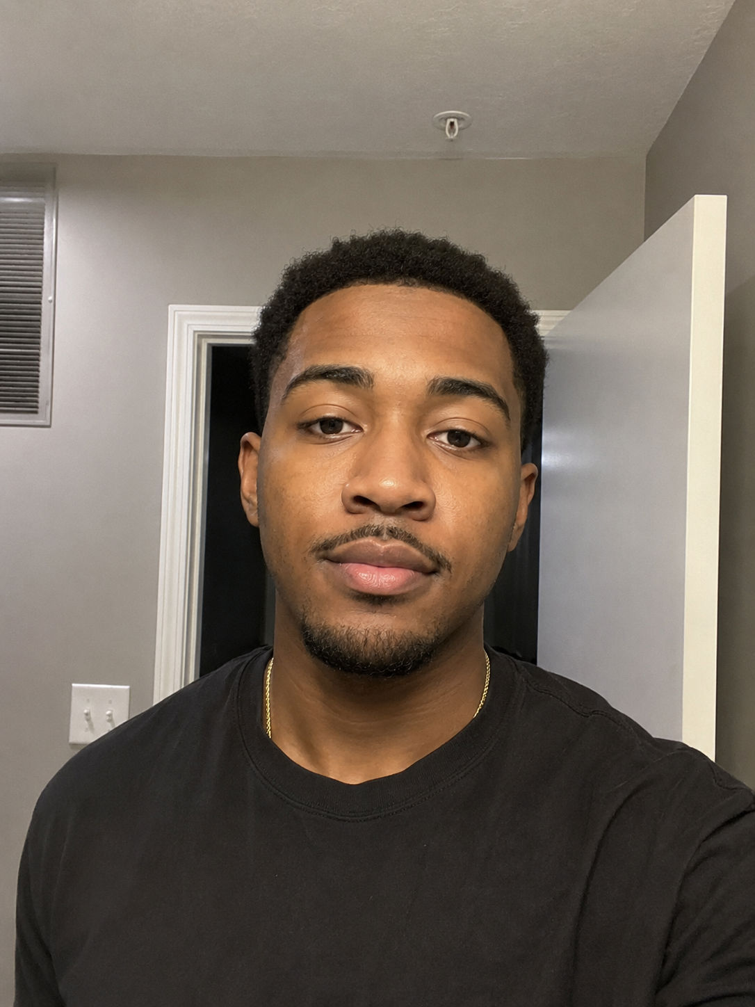 A young man with short curly hair and a mustache taking a selfie indoors, wearing a black T-shirt and a gold chain, with a white door and gray walls in the background.