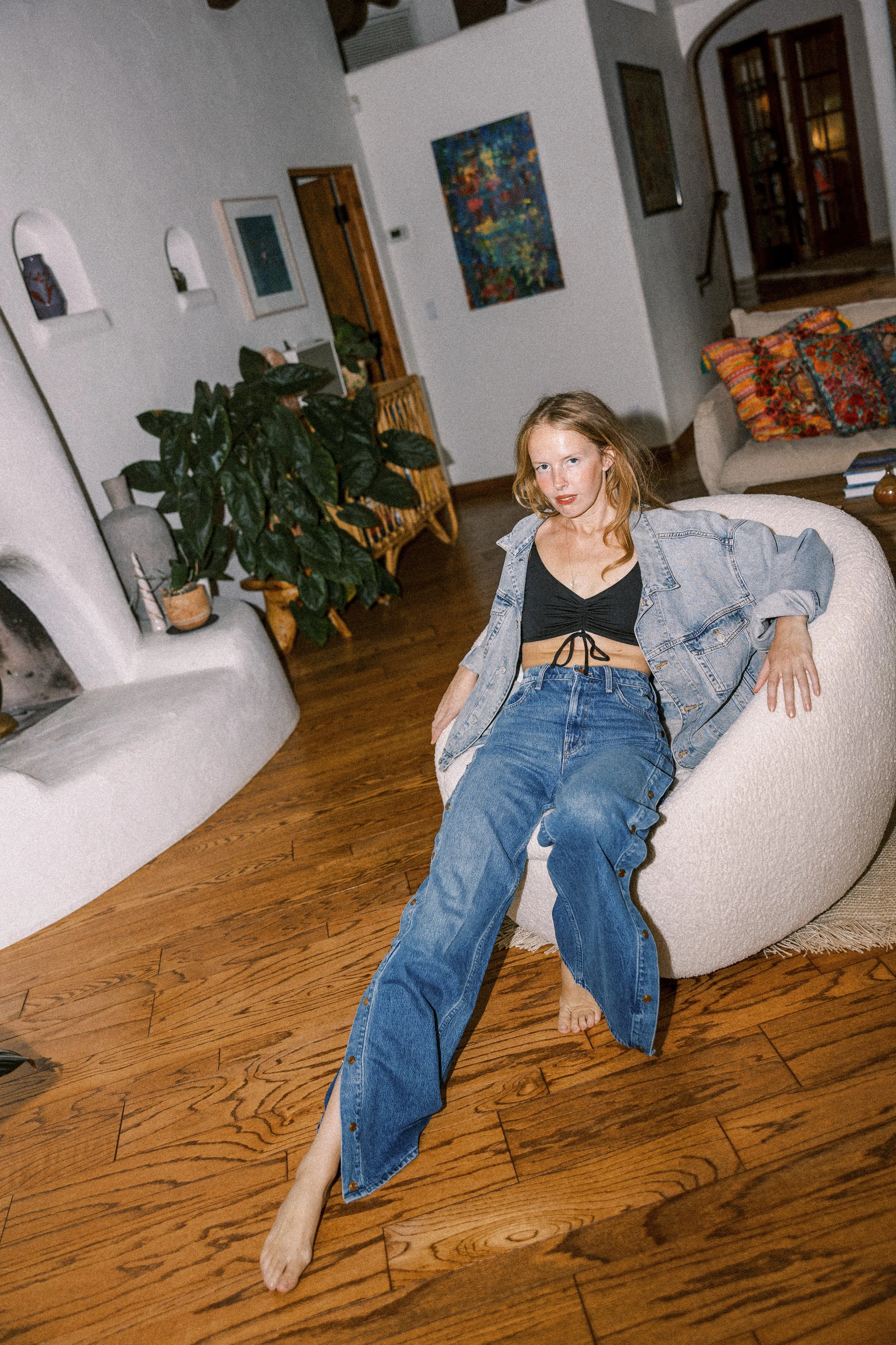 Studio owner Tara Livdeep with reddish hair sitting on a beige armchair in a living room, wearing a black top, a denim jacket, and baggy jeans. She is barefoot, with her feet on the wooden floor, and looks directly at the camera.