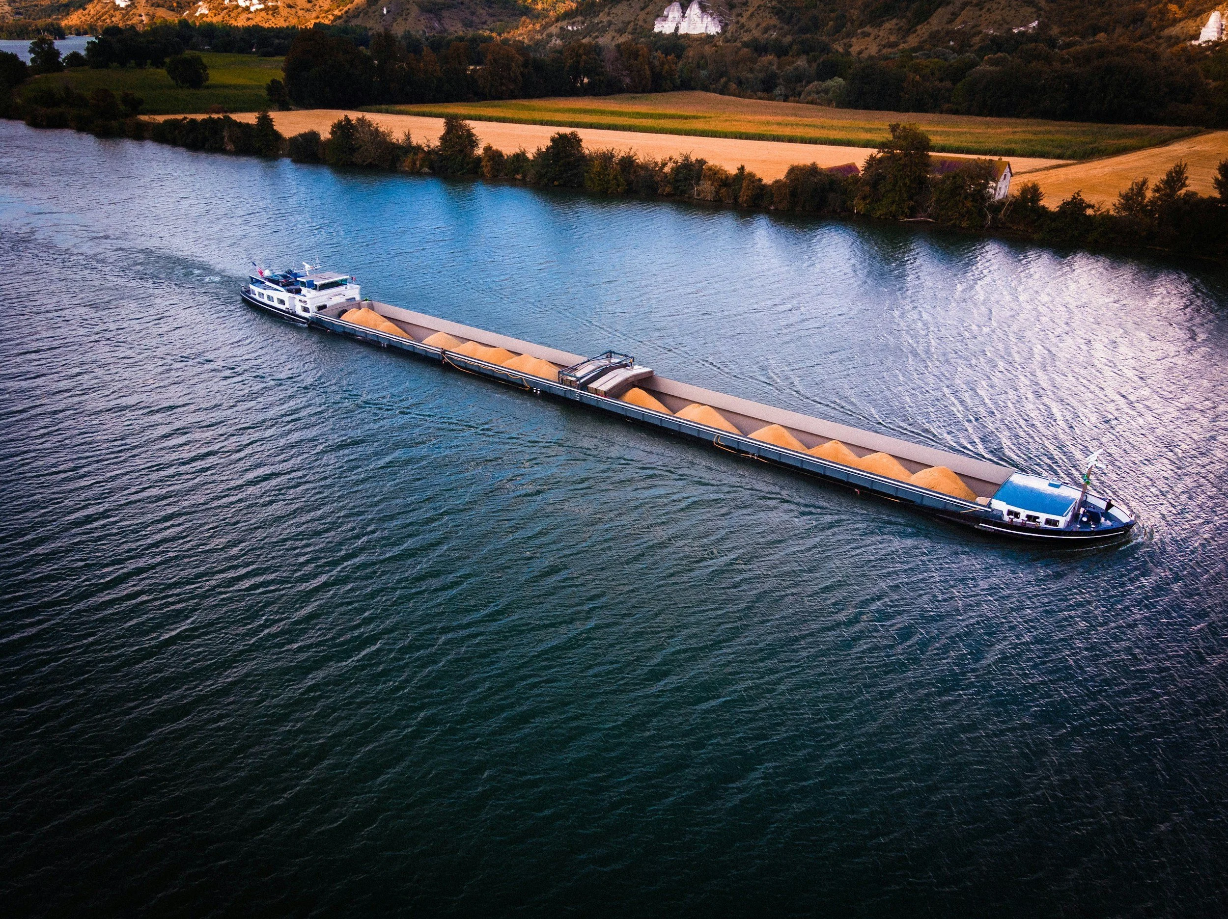 A large cargo ship with beige material on its deck is moving through a wide river or lake, surrounded by lush green and brown hills.