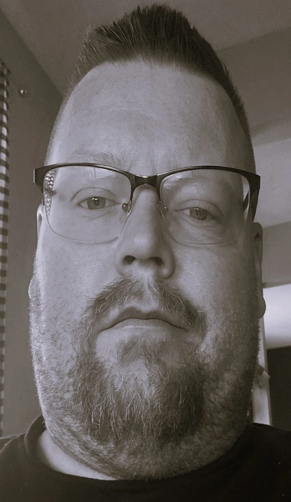 Close-up black and white portrait of a man with glasses, a beard, and a mustache, looking directly at the camera.