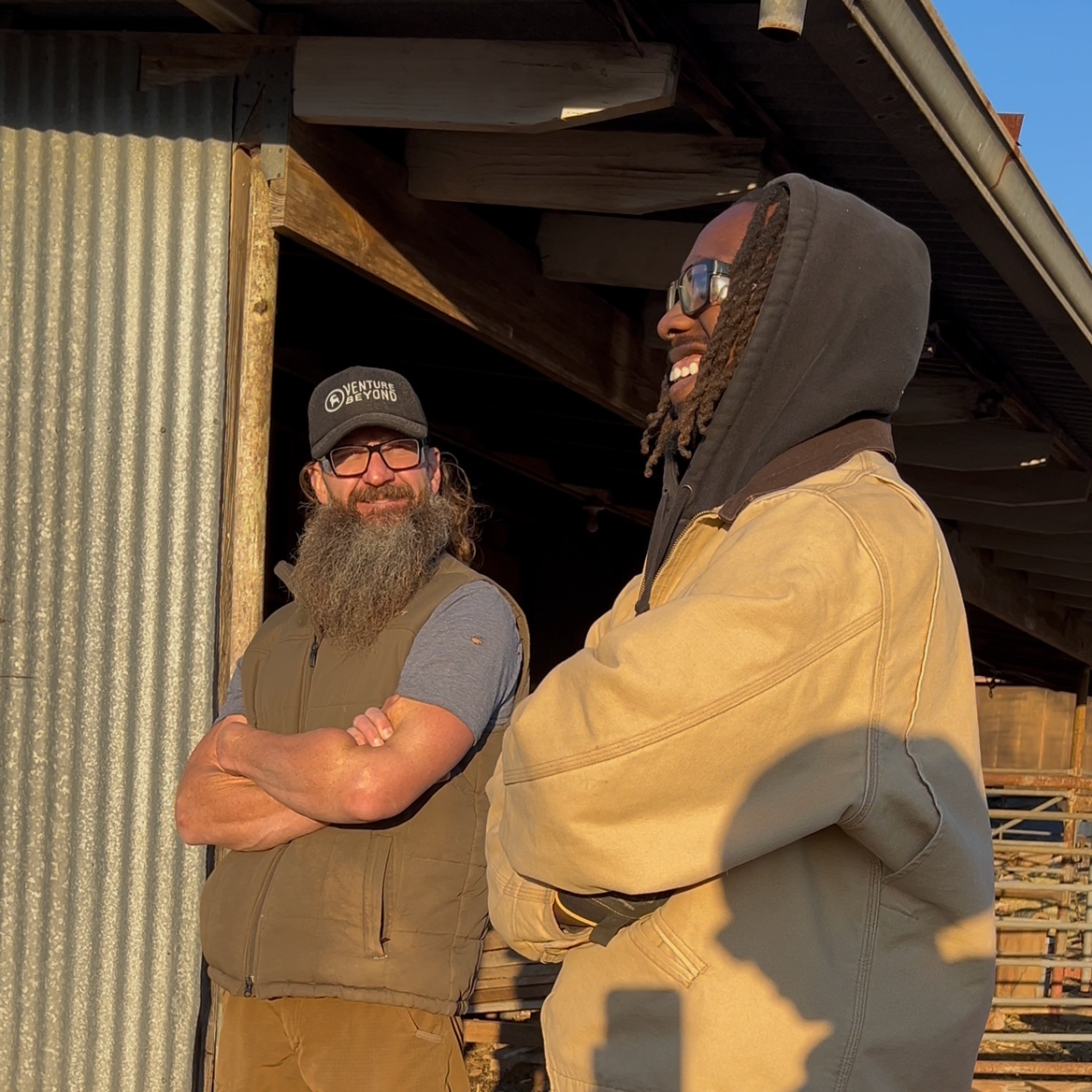 Two men standing outside near a rustic building, smiling and talking. One has a beard, glasses, a black cap, and a tan vest. The other has dreadlocks, glasses, and is wearing a yellow jacket with a black hoodie underneath. Sunlight is shining on them.
