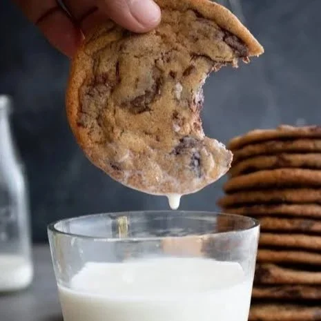 A hand holding a chocolate chip cookie above a glass of milk, with a bite taken out of the cookie and milk dripping from it.