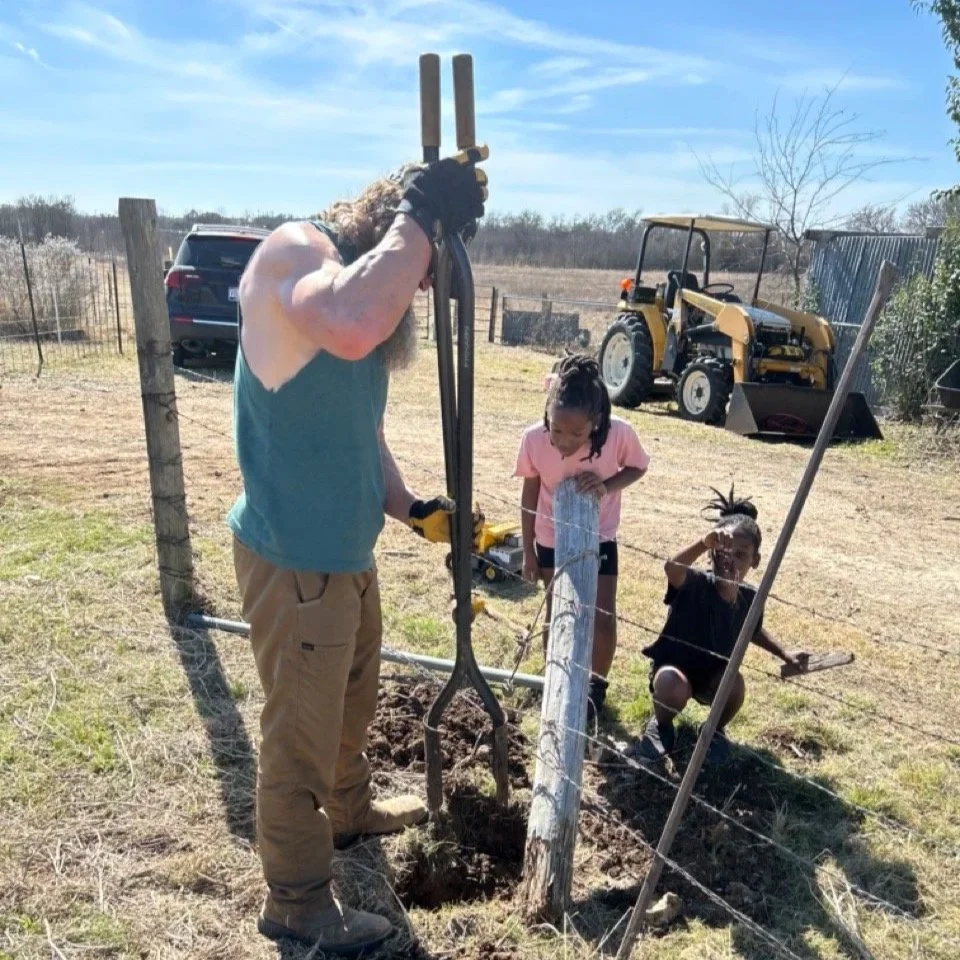 A man with long curly hair and a beard planting a small tree with three kids in a field. The man is holding a large shovel, while the kids observe and assist. There is a yellow tractor and a parked vehicle in the background, along with a fenced area and a blue sky.