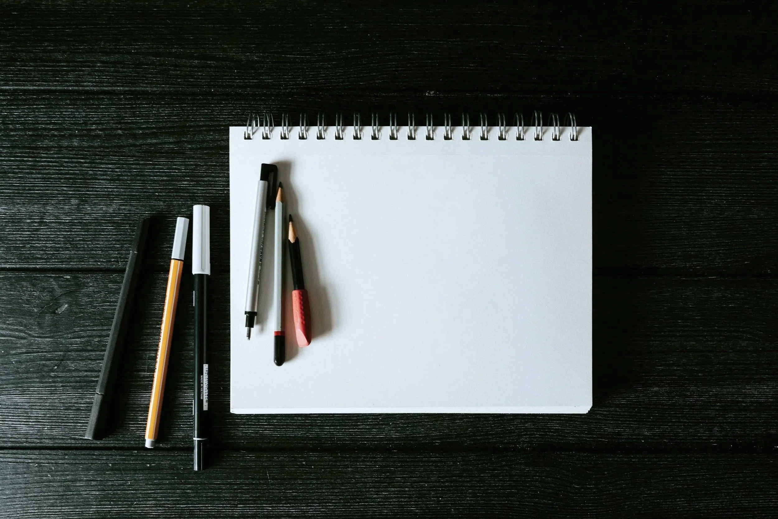 Blank spiral notebook with pencils, pens, and markers on a black wooden desk