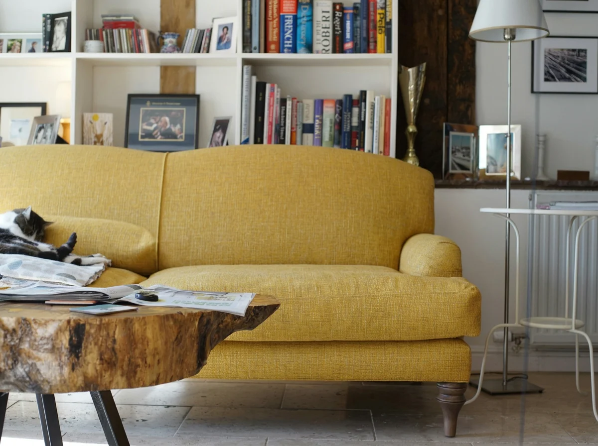 Black-and-white cat reclining on a mustard-yellow sofa beside a rustic wooden coffee table in a cozy living room.