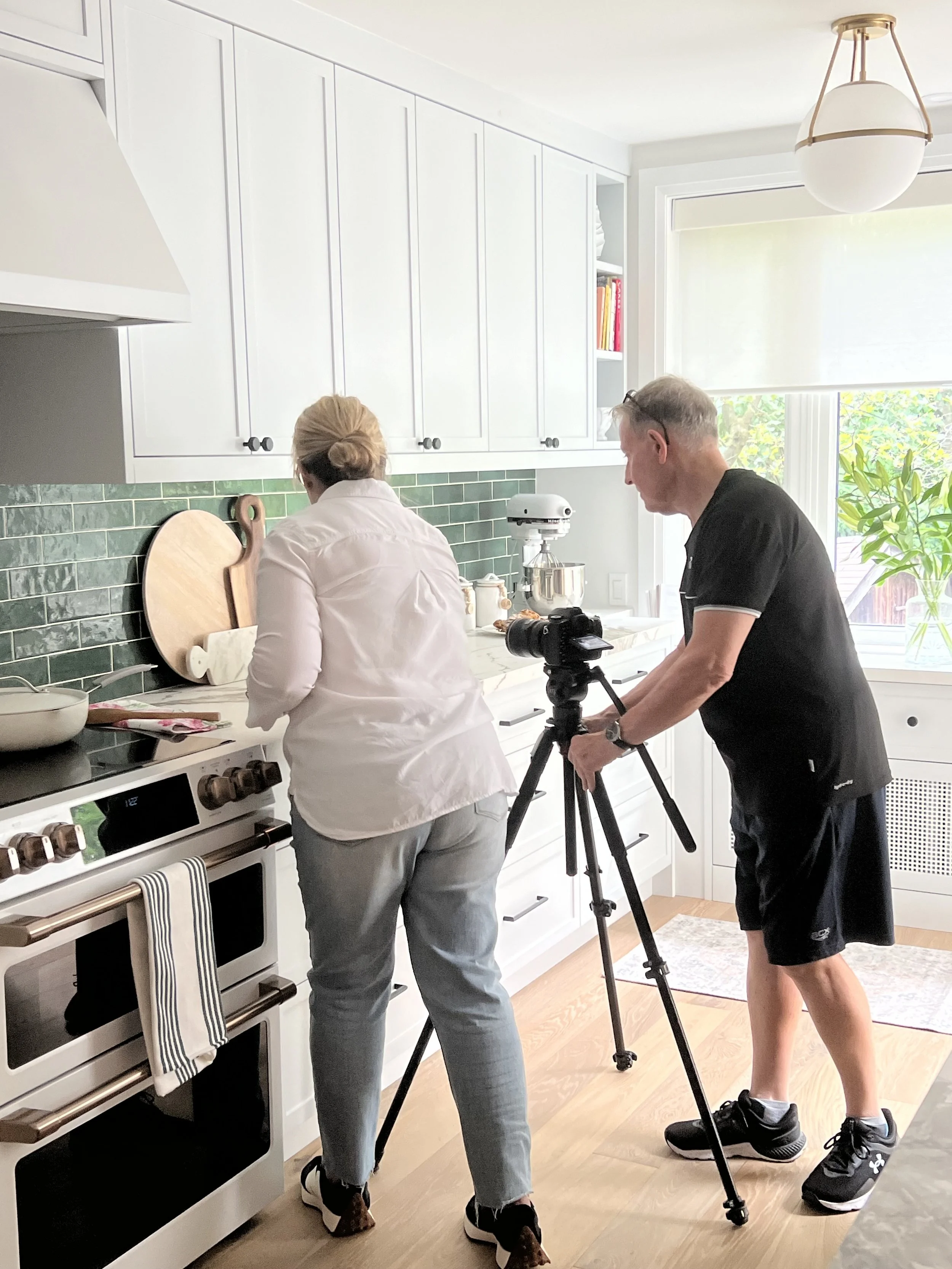 Two people filming styling photographs in a bright modern kitchen with a camera on a tripod and stand mixer.