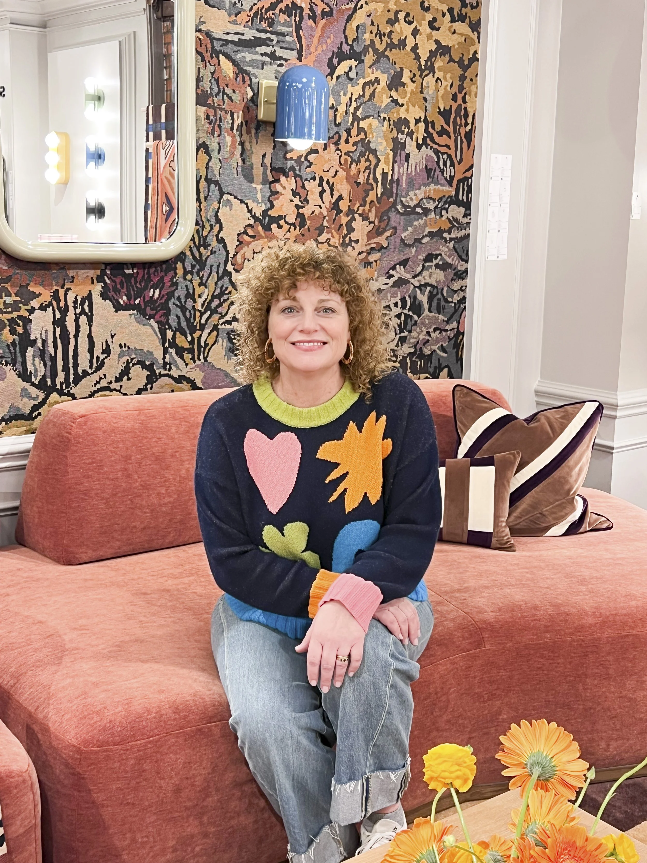 Smiling Miriam seated on a coral sofa in a colorful living room with abstract wallpaper, blue wall lamp, and orange flowers.