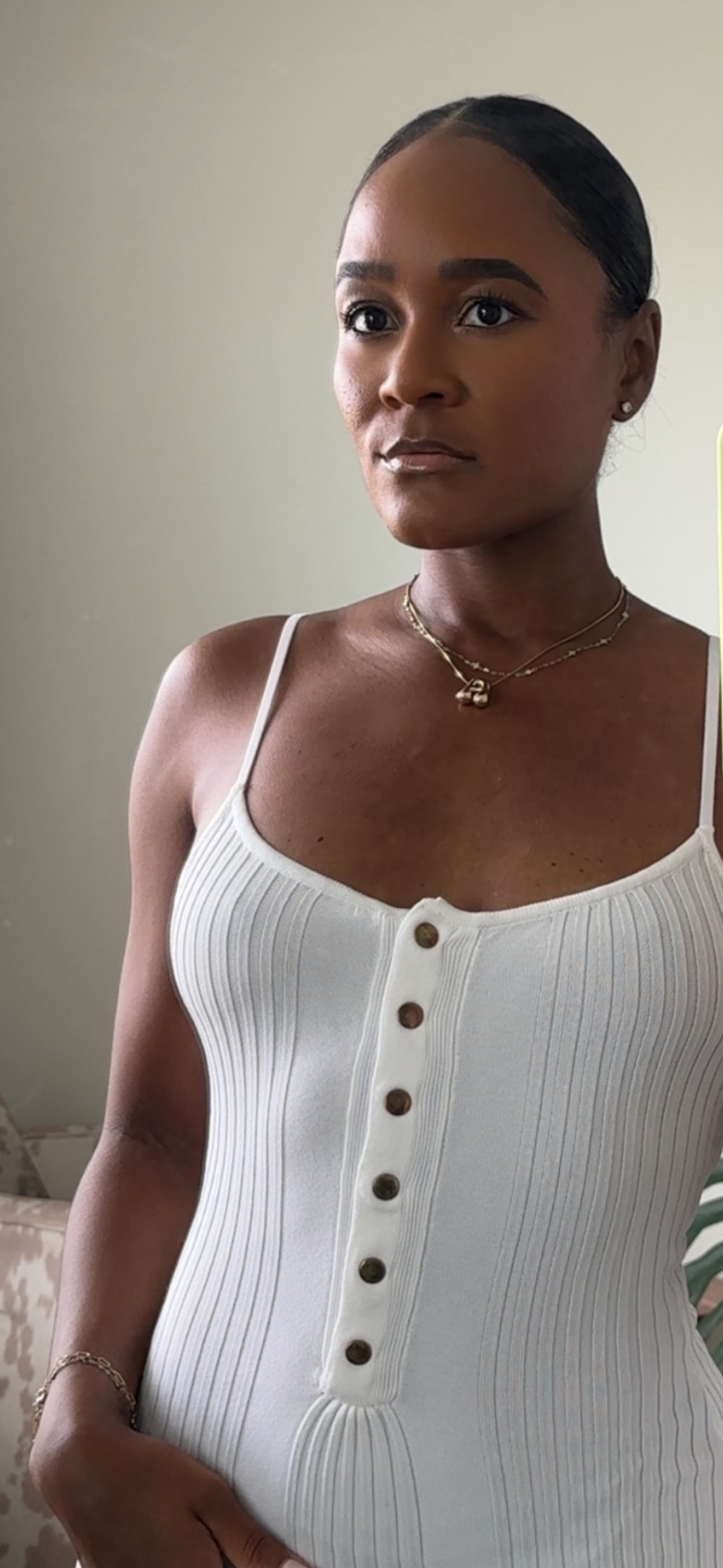 A woman with dark hair slicked back, wearing a sleeveless white ribbed button-up top, and gold jewelry including a necklace and bracelet, standing against a plain wall.