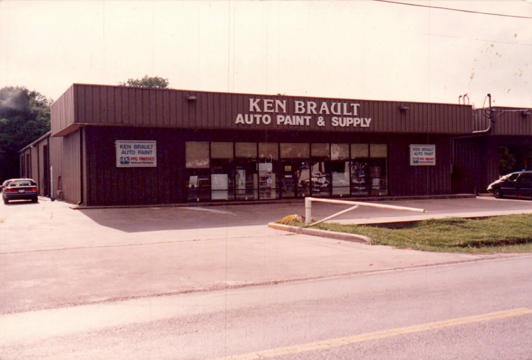 Photo of a vintage storefront with a sign reading 'Ken Brault Auto Paint & Supply.' The building has large glass windows and is painted dark. There are a couple of cars parked in front, with a small landscaped area and a parking lot.
