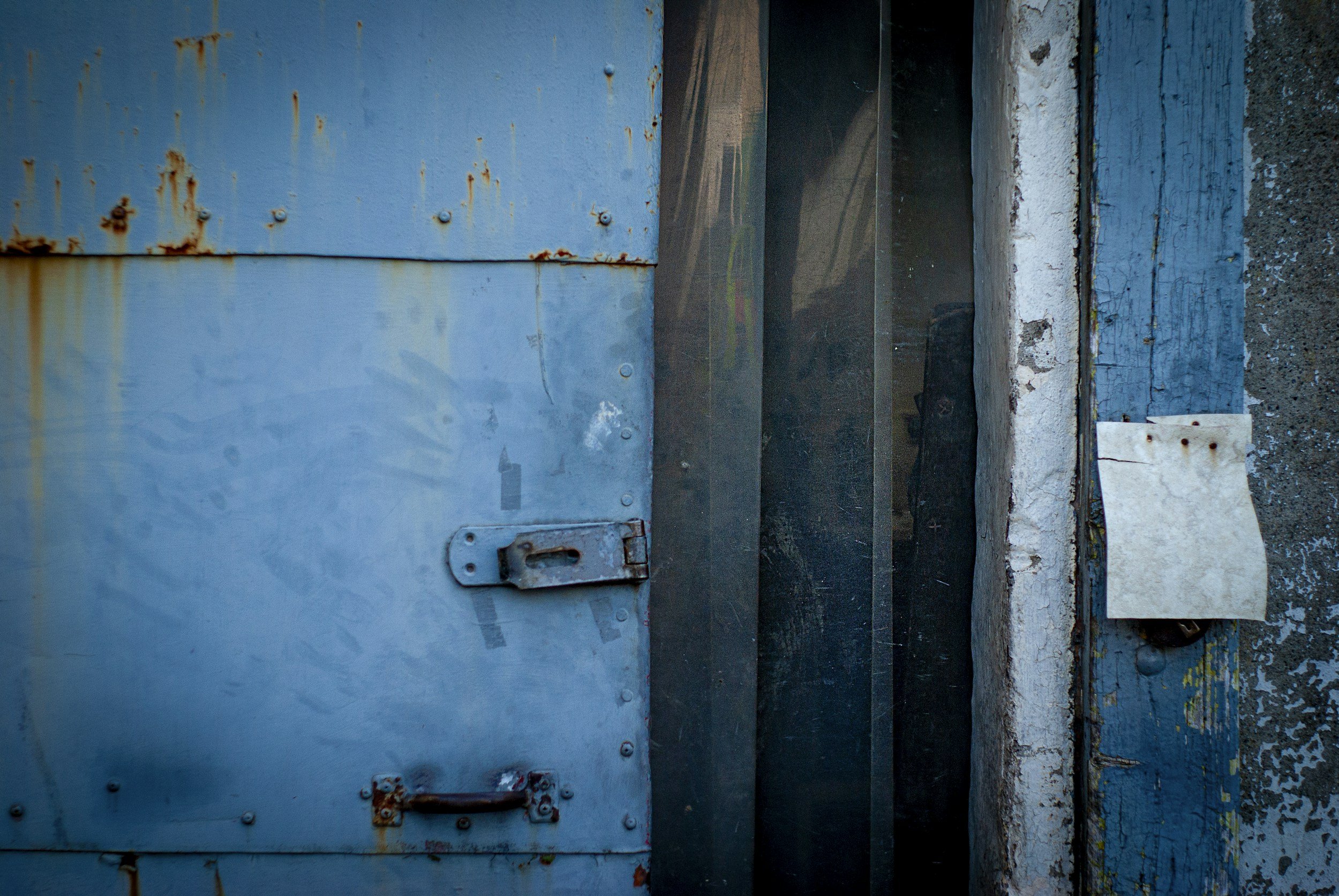 Close-up of a weathered blue metal structure with rust spots, bolts, and hinges, adjacent to a concrete wall and a piece of wood in a gritty urban setting.