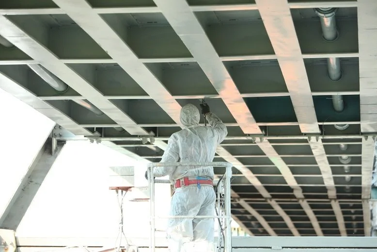 Worker in protective gear painting or inspecting an underside of a bridge or large structure.