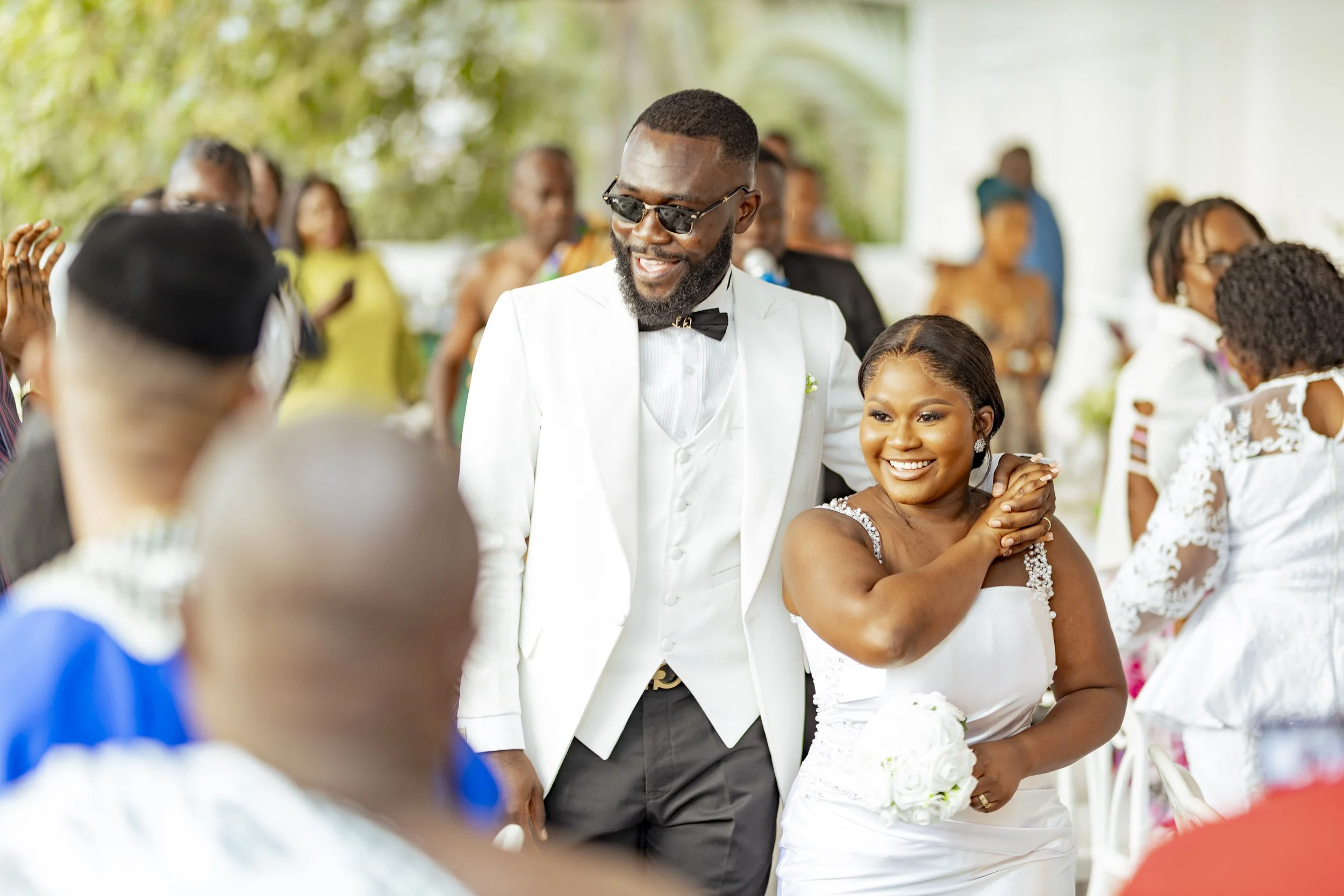 A groom and bride at a wedding reception, with the groom wearing sunglasses and a white tuxedo jacket, and the bride in a white wedding dress holding a bouquet, smiling and standing closely together.
