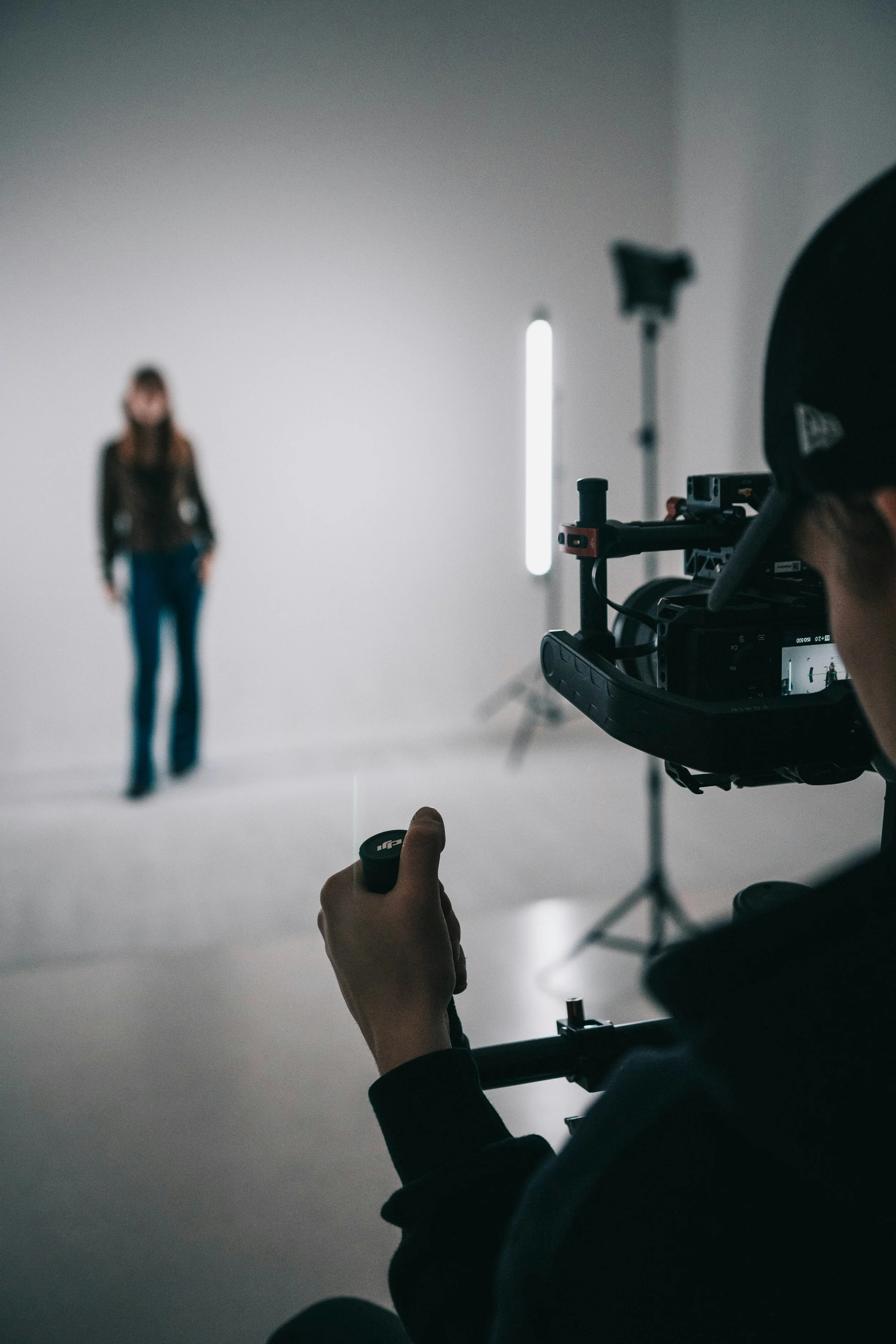 A person holding a remote control in front of a professional video camera in a studio, with a woman standing in the background. The studio has a plain white backdrop, and studio lights are visible.