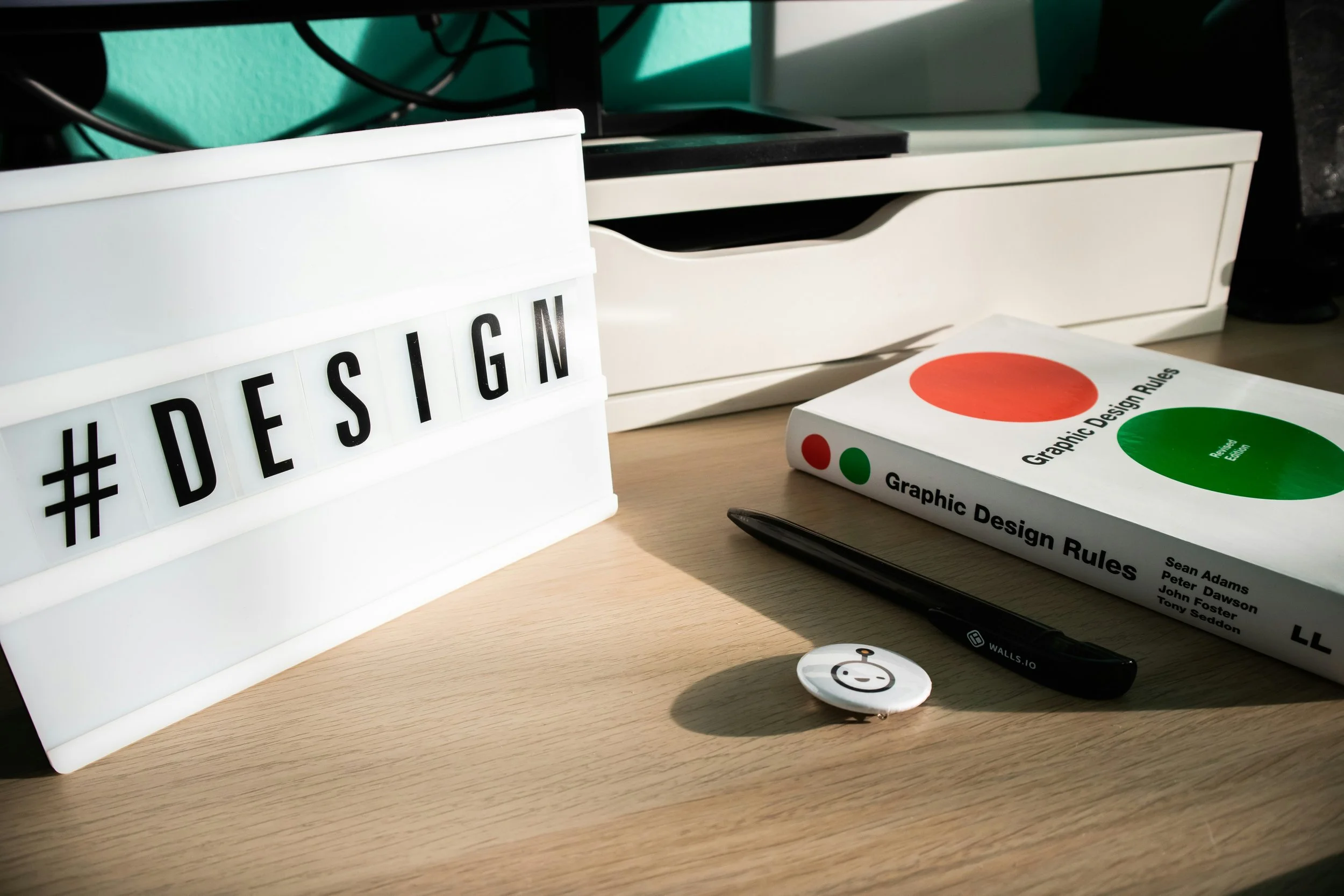 A desk with a white lightbox displaying '#DESIGN', a book titled 'Graphic Design Rules', a black pen, a small round smiley face pin, and various items including a printer and cables in the background.