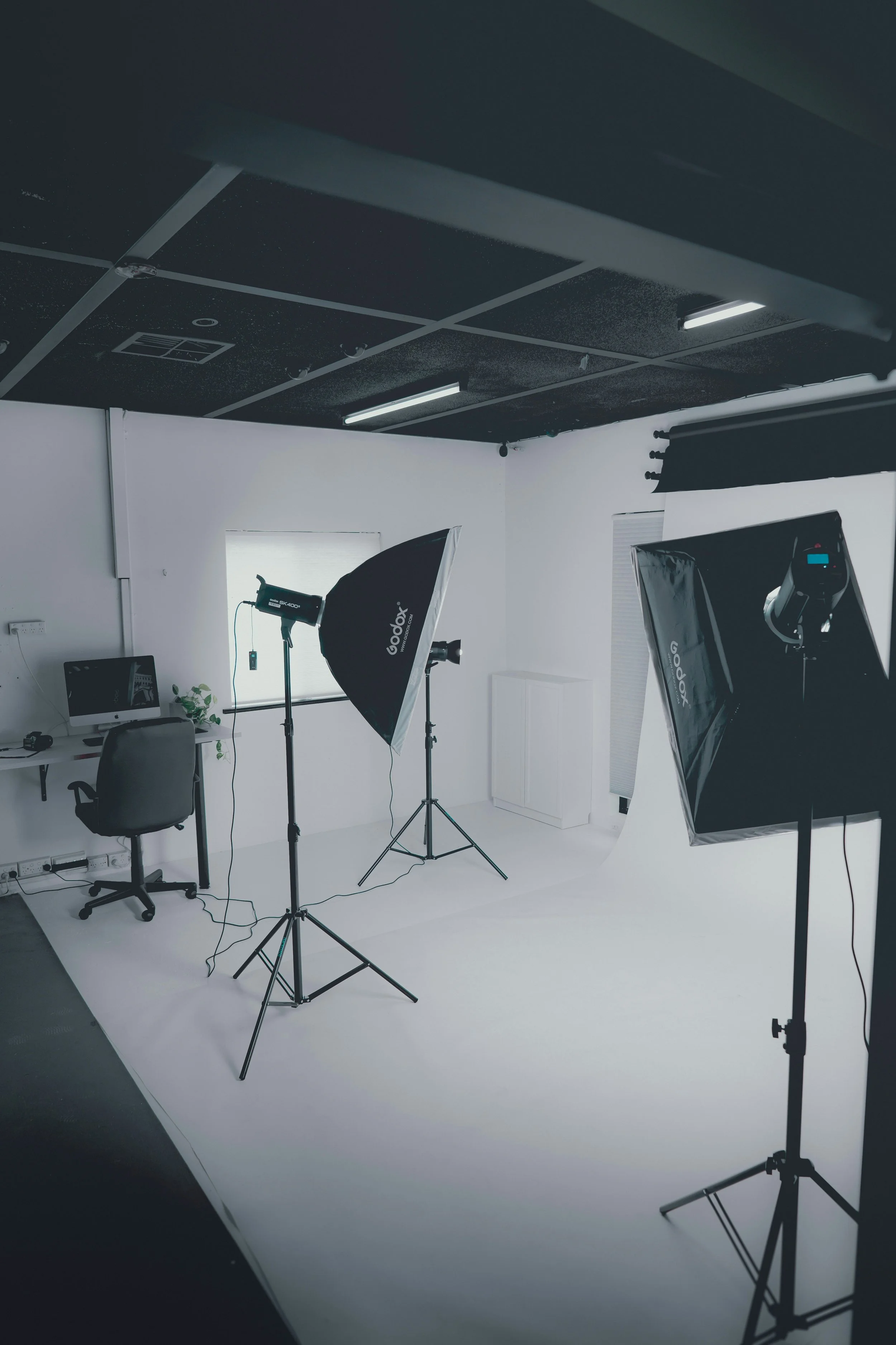 A photography studio setup with lighting equipment, a white seamless background, and a computer desk with an iMac and headphones, indicating a professional photo shoot environment.