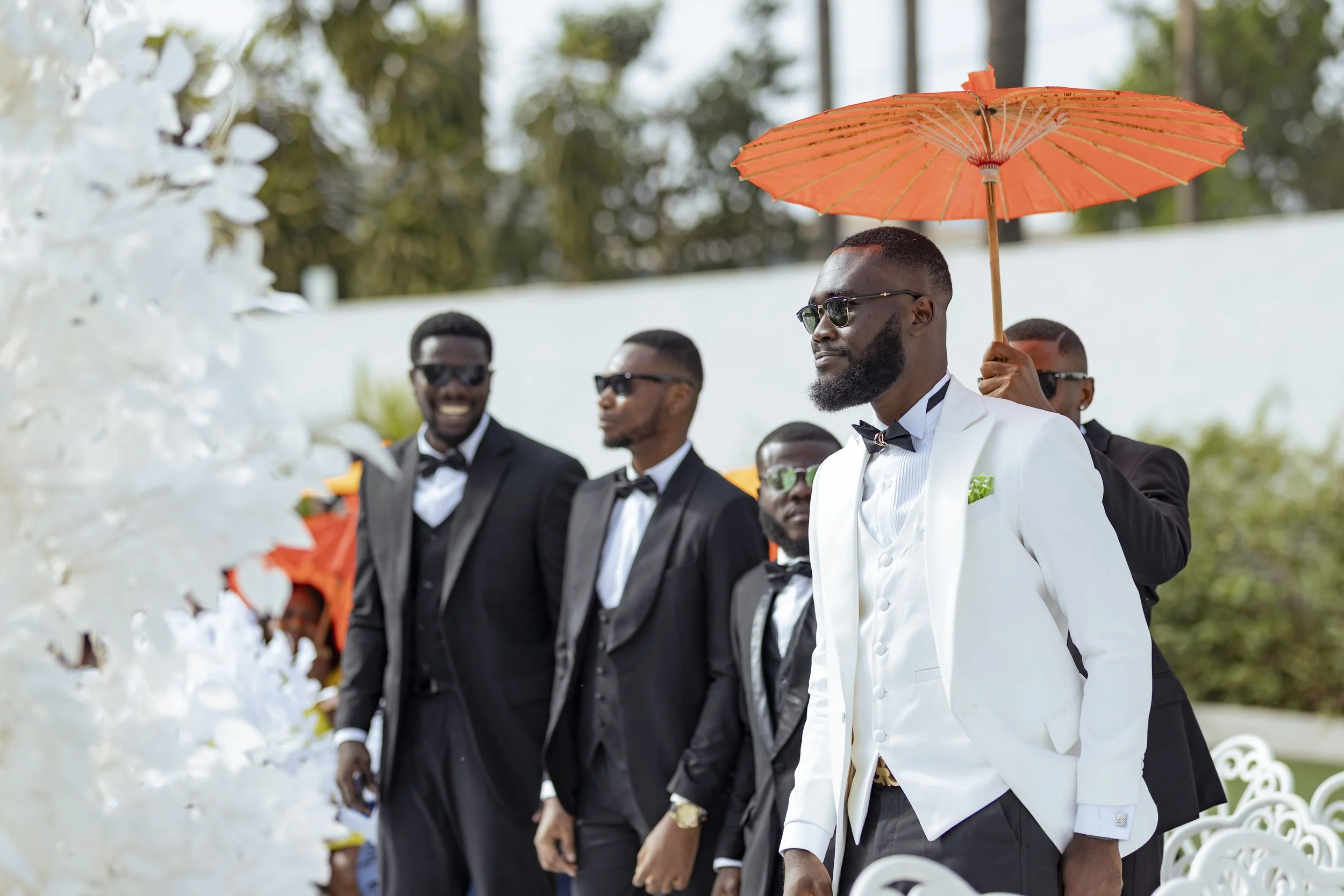 A man in a white tuxedo with a bow tie holding an orange parasol while standing among a group of men in black tuxedos at an outdoor wedding ceremony.