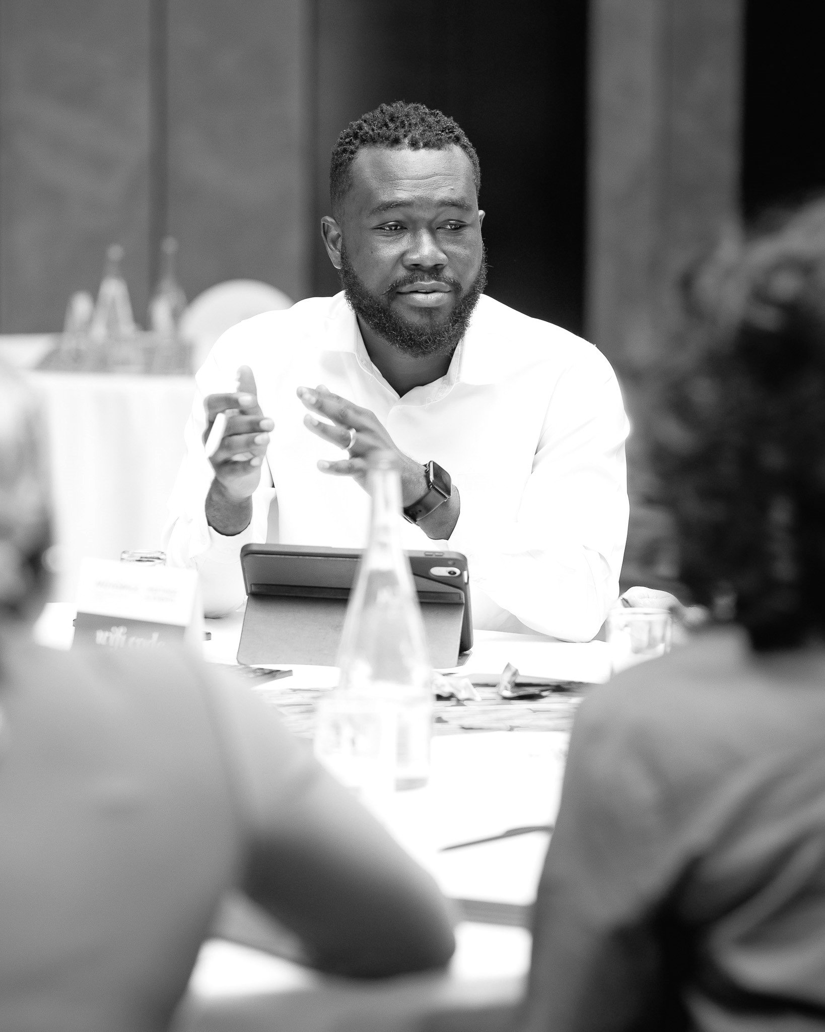 Black and white photo of a man with a beard and short curly hair, sitting at a table with a serious or contemplative expression, with blurred objects on the table and background.