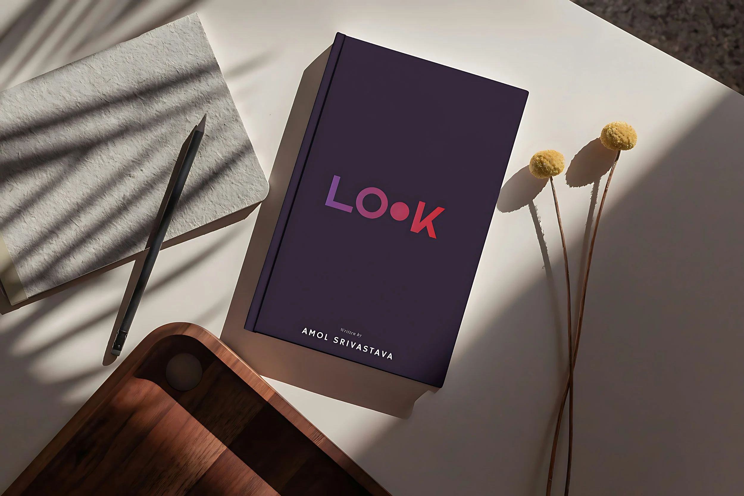 A book titled 'Look' by Amol Srivastava on a white table, with two yellow billy button flowers and a black pen on a notepad, all illuminated by sunlight with shadows.