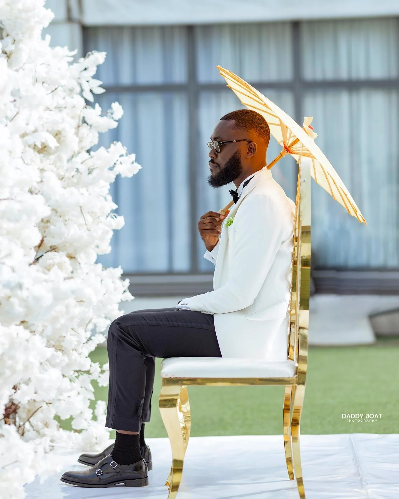 Man dressed in a white tuxedo with a black bow tie and sunglasses, sitting on a gold chair, holding a white parasol, next to a white flowering tree during daytime.
