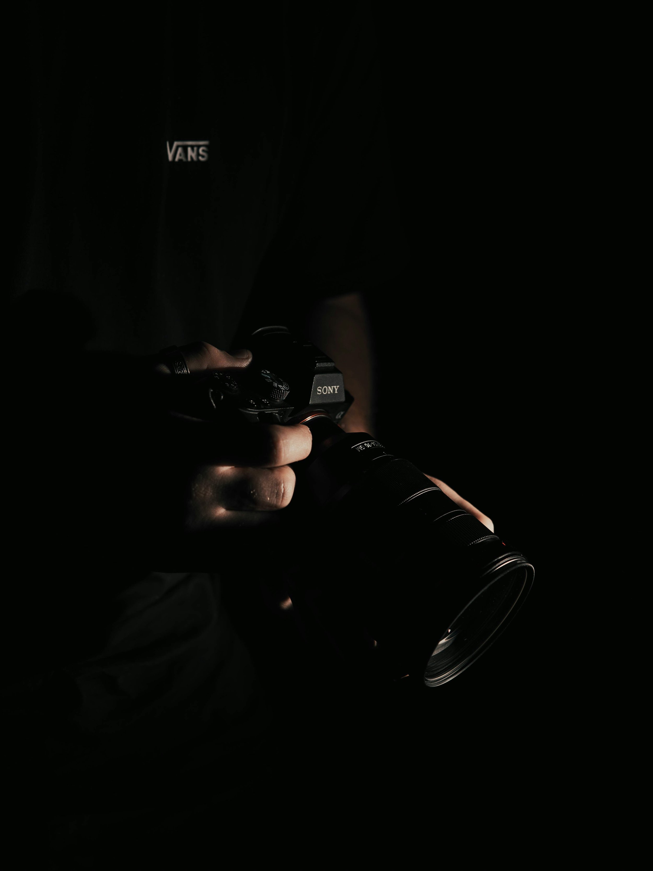 Person holding a Sony camera with a large lens in a dark environment.