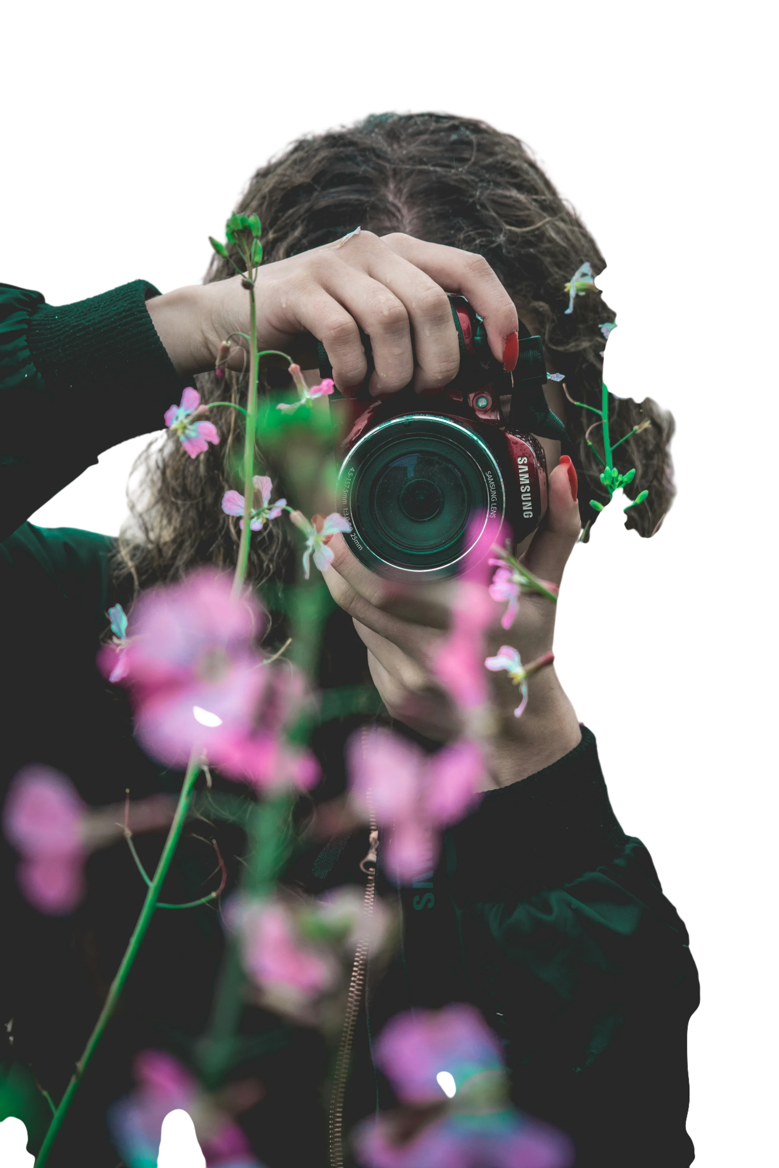 A person with curly hair taking a photo with a red Samsung camera, surrounded by pink flowers.