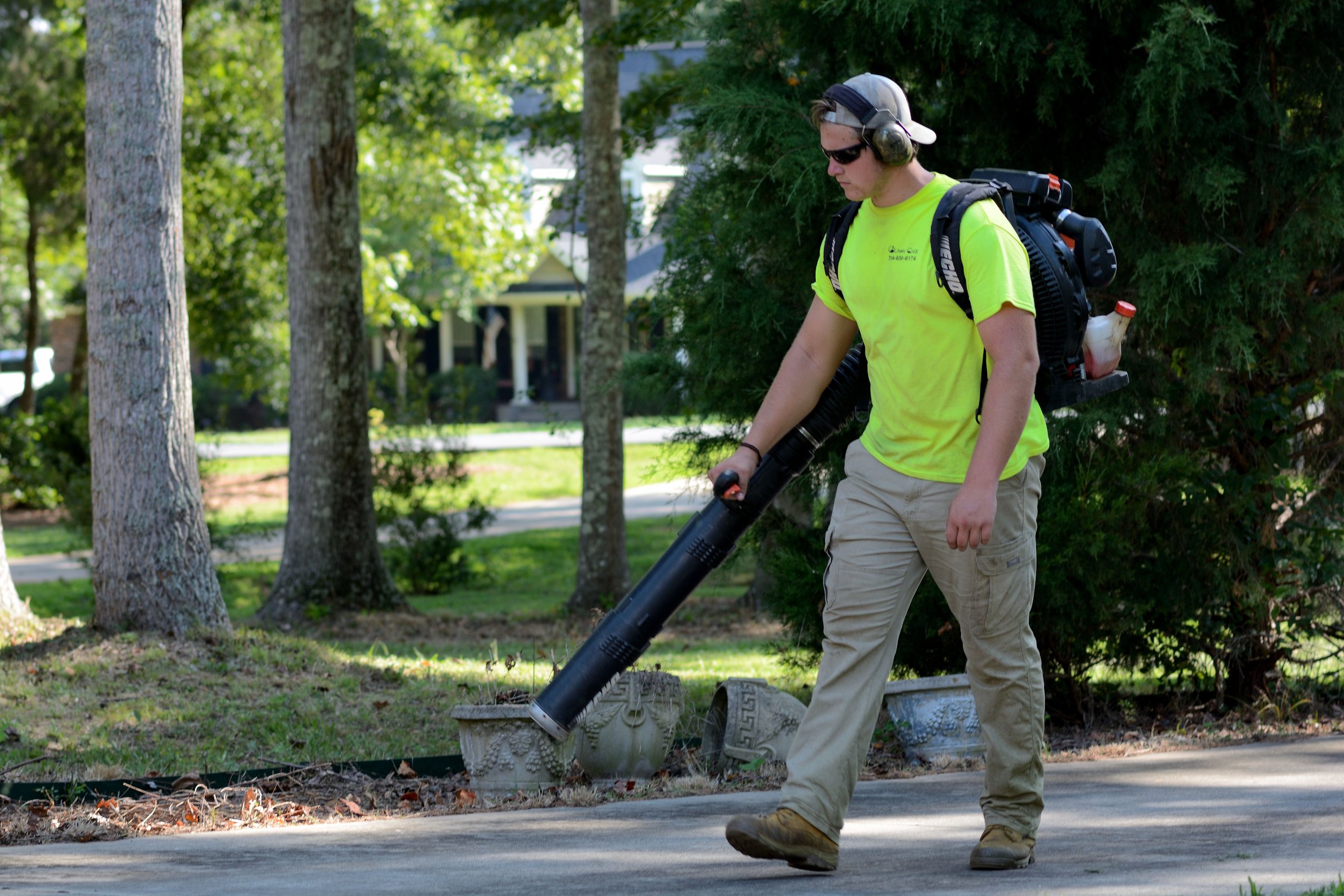 A man in a neon yellow shirt, khaki pants, sunglasses, and a cap is using a leaf blower to clear fallen leaves on a sidewalk in a residential neighborhood with trees and houses in the background.