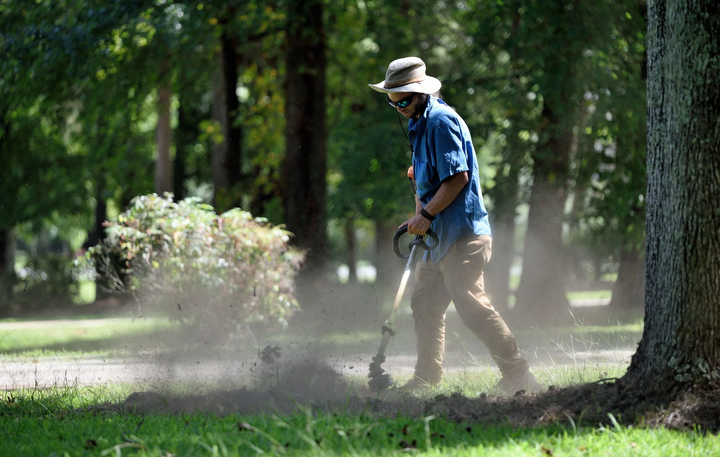 A man wearing a wide-brimmed hat, sunglasses, a blue shirt, and brown pants is trimming bushes in a park with a string trimmer, creating dust and debris as he works.