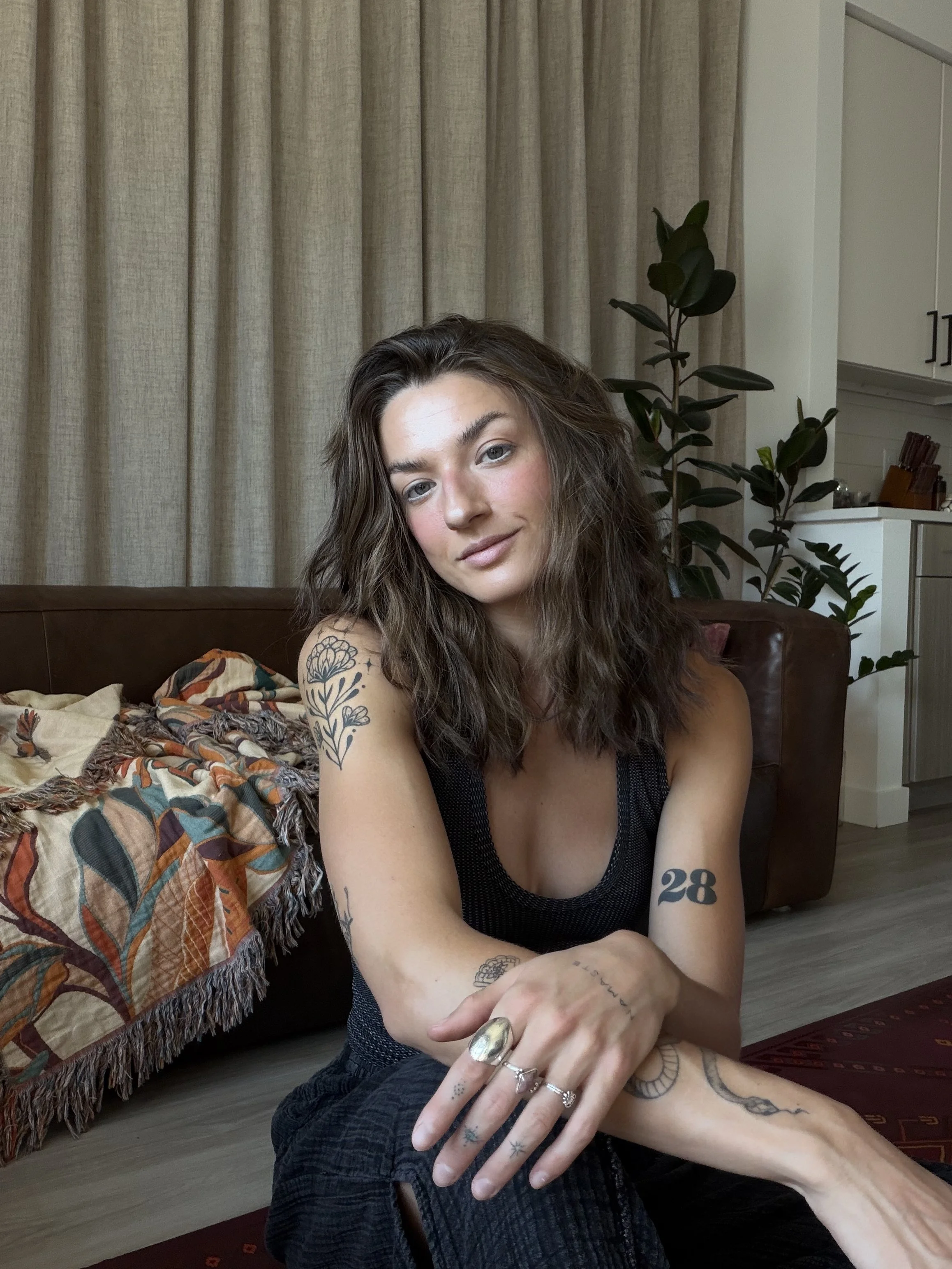 A young woman with wavy brown hair and tattoos on her arms sitting on the floor in a room with beige curtains, plants, and a white cabinet, wearing a black tank top and black shorts.