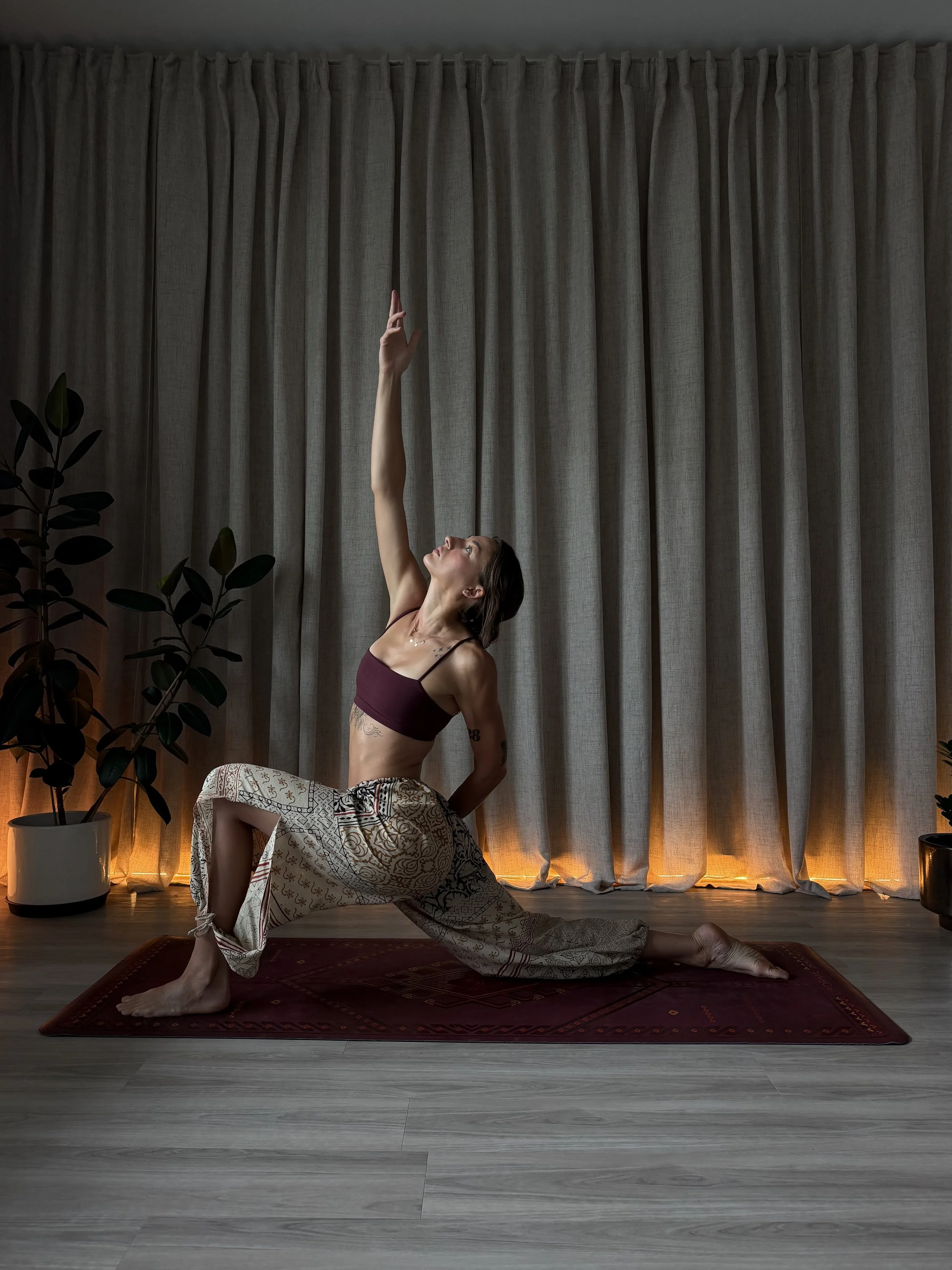 A woman practicing yoga indoors, performing a low lunge pose on a red patterned rug with a burgundy top and patterned pants. She is reaching upward with one arm, with long curtains behind her, potted plants to her side, and soft lighting creating a peaceful atmosphere.