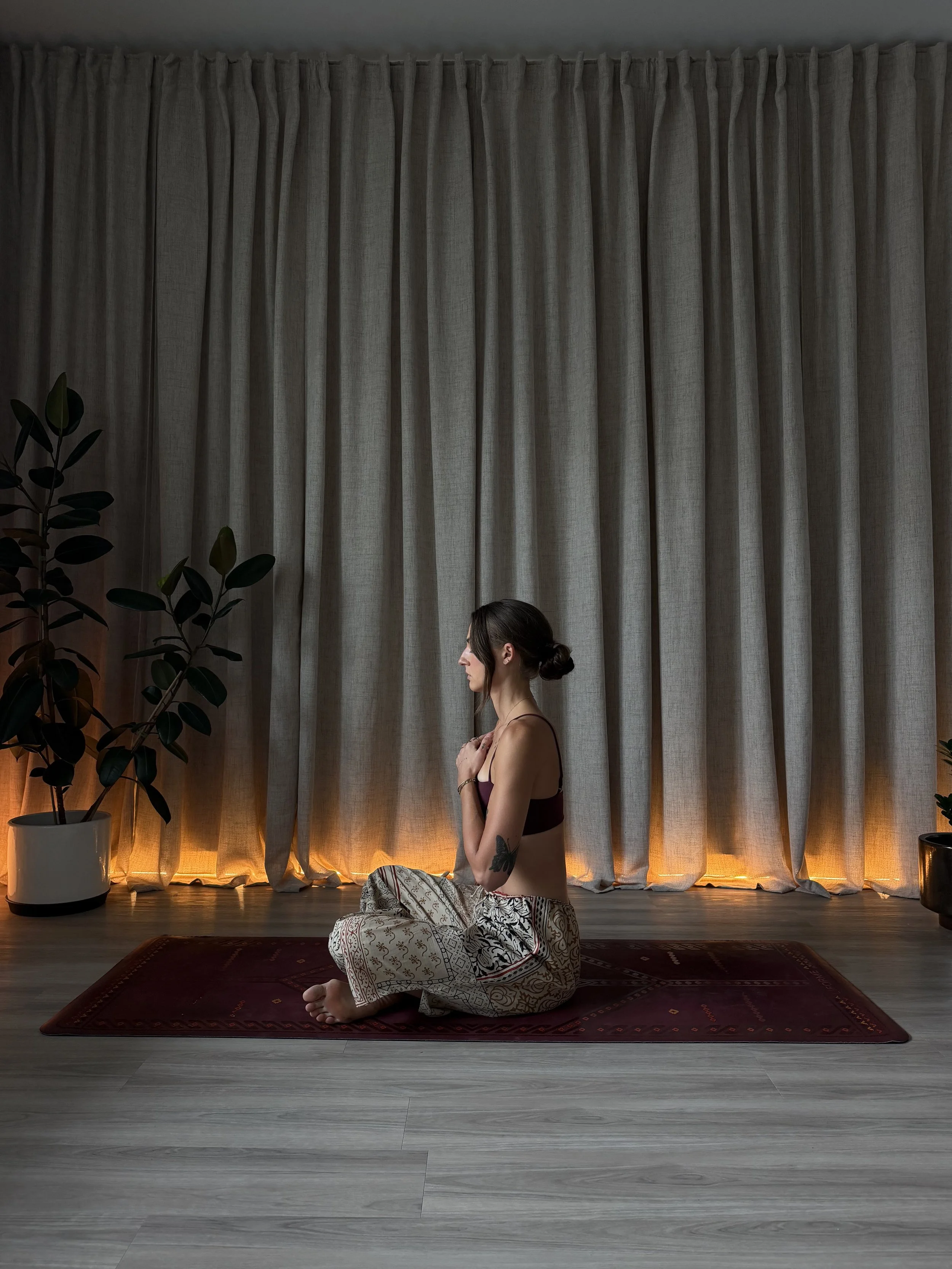 A woman practicing yoga or meditation in a serene indoor setting. She is sitting cross-legged on a patterned rug, with her eyes closed and hands resting gently on her chest. Behind her, there are large beige curtains, tall leafy potted plants on either side, and soft warm lighting creating a calming atmosphere.