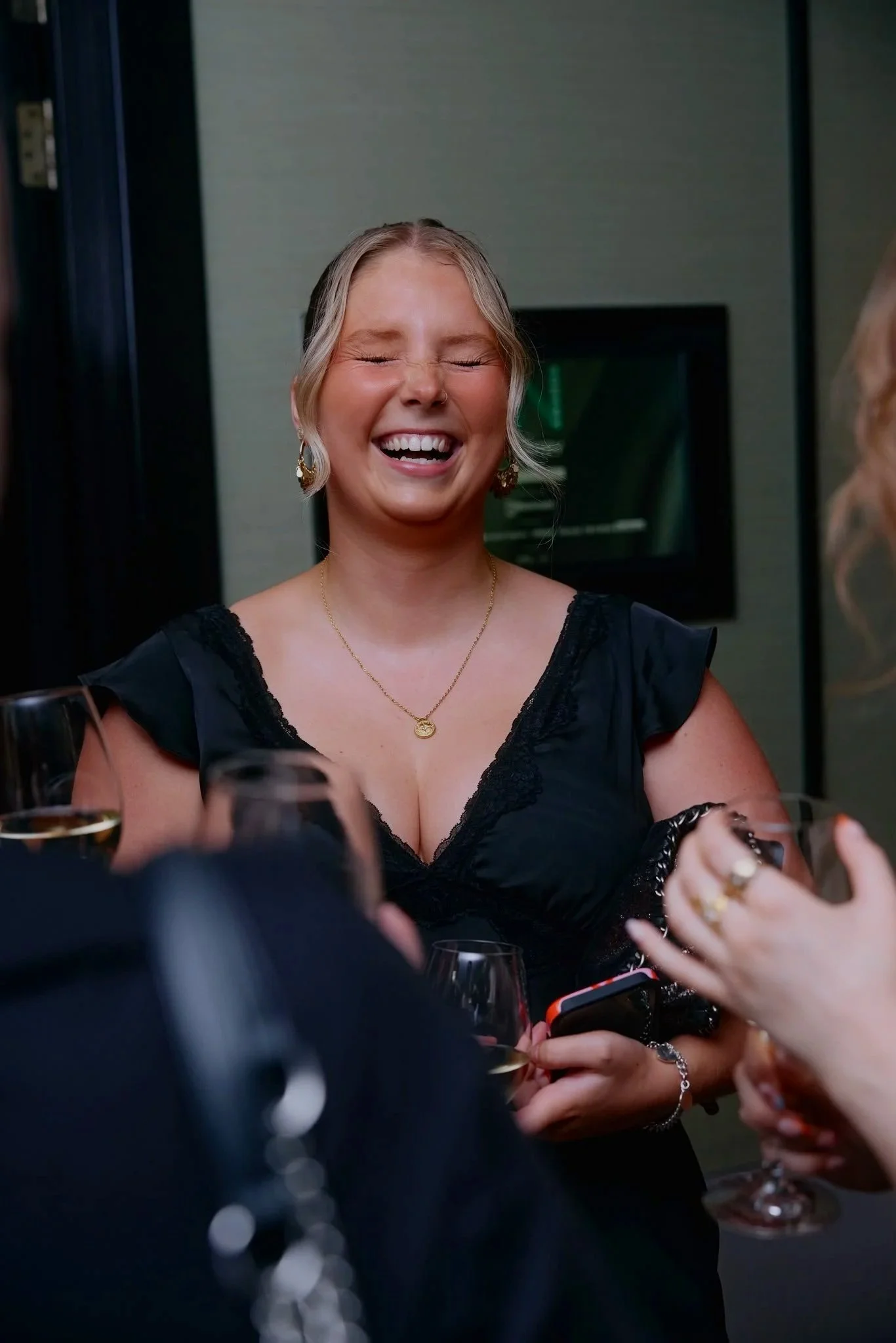 A woman with blonde hair wearing a black dress with lace trim, gold jewelry, and earrings is smiling with her eyes closed at a social gathering, holding a glass of wine.