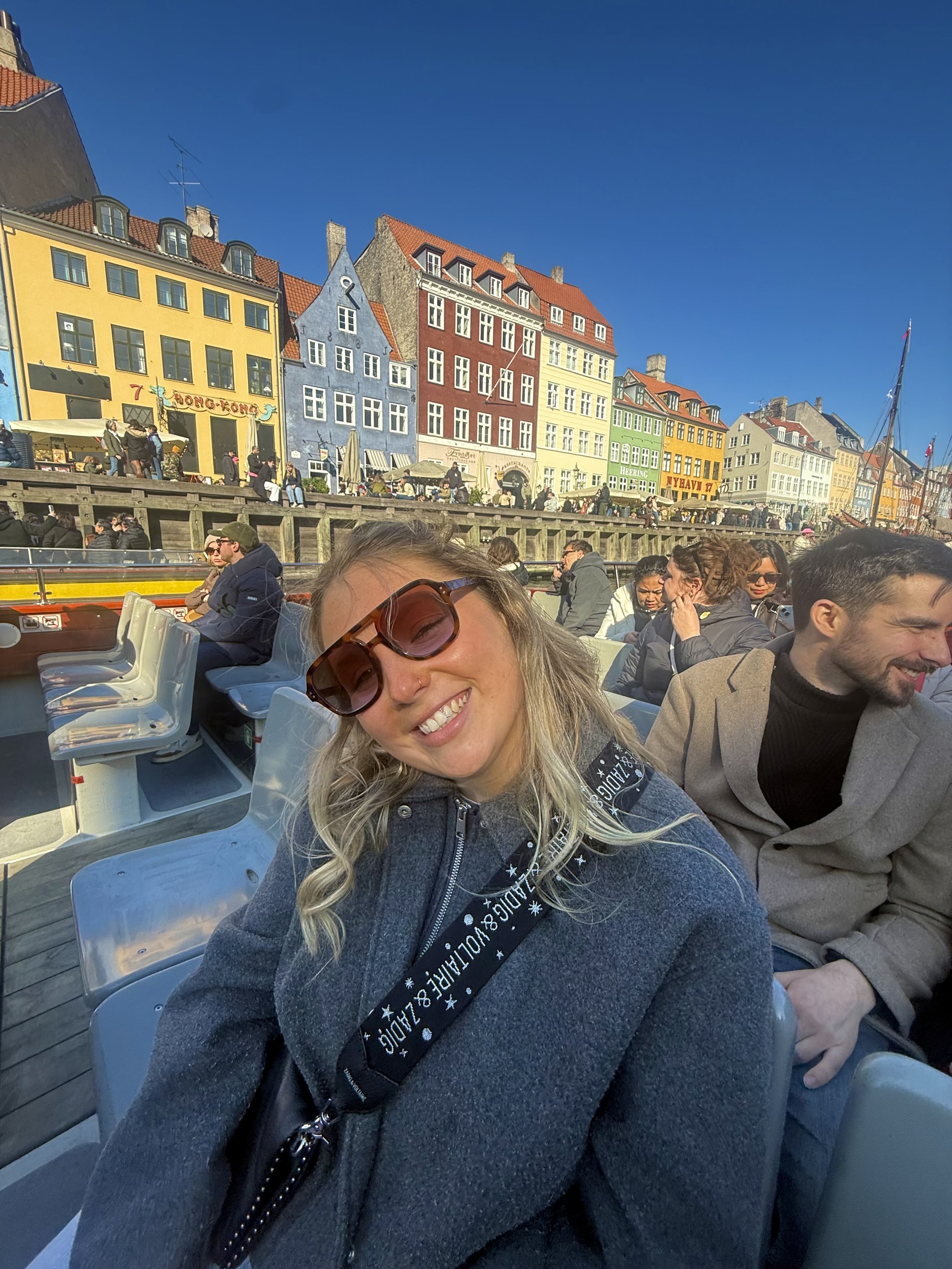A woman wearing sunglasses and a dark sweater smiling on a boat, with colorful buildings and other passengers in the background.
