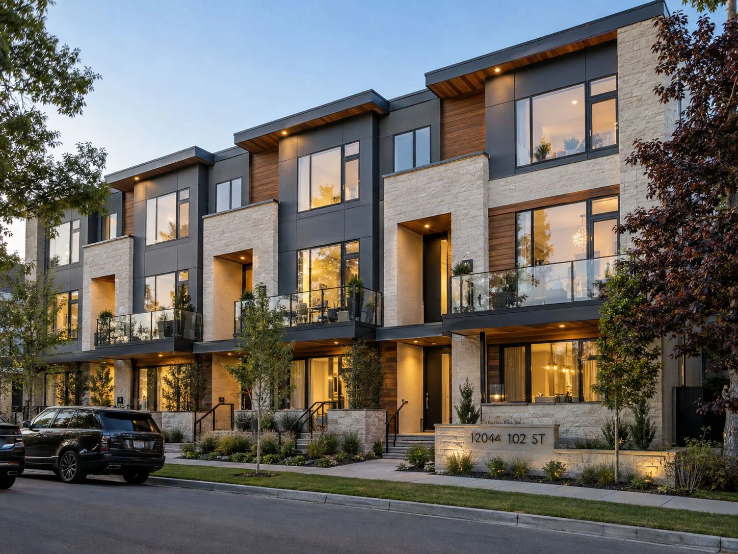 Modern multi-story apartment building with large windows, balconies with glass railings, and landscaped front yard. The building has a mix of stone, wood, and metal siding, illuminated by warm exterior lighting at dusk.