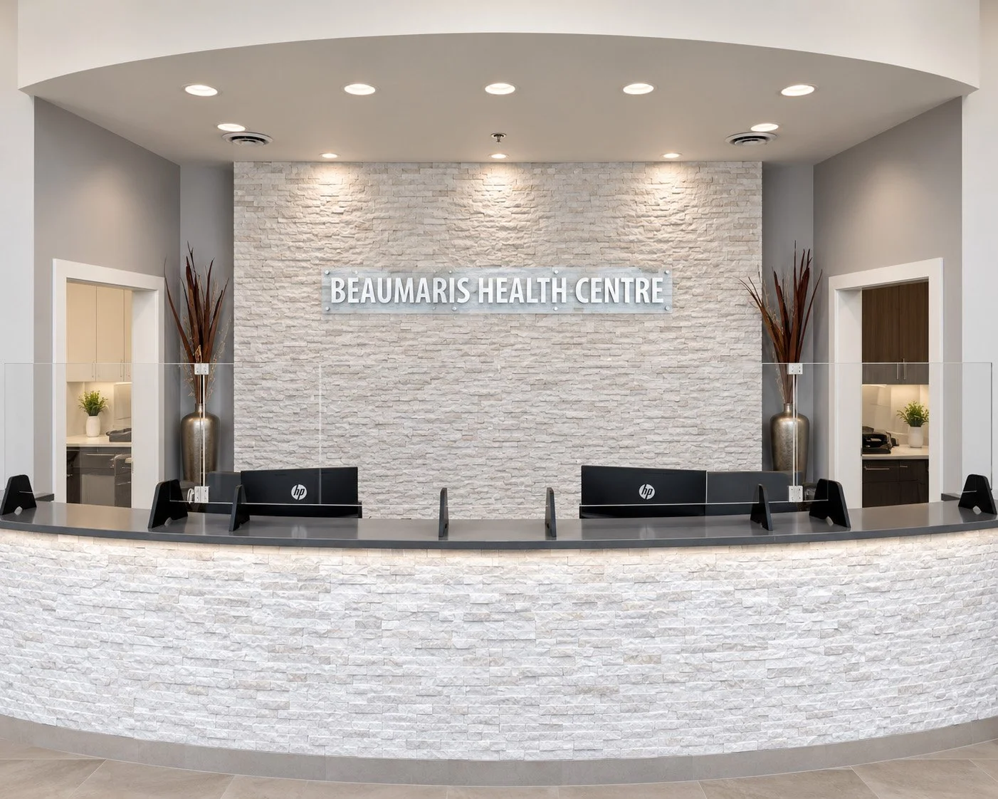 Reception area of Beaumaris Health Centre with a white stone textured wall, a sign, two reception desks with computers, and decorative vases with tall branches.