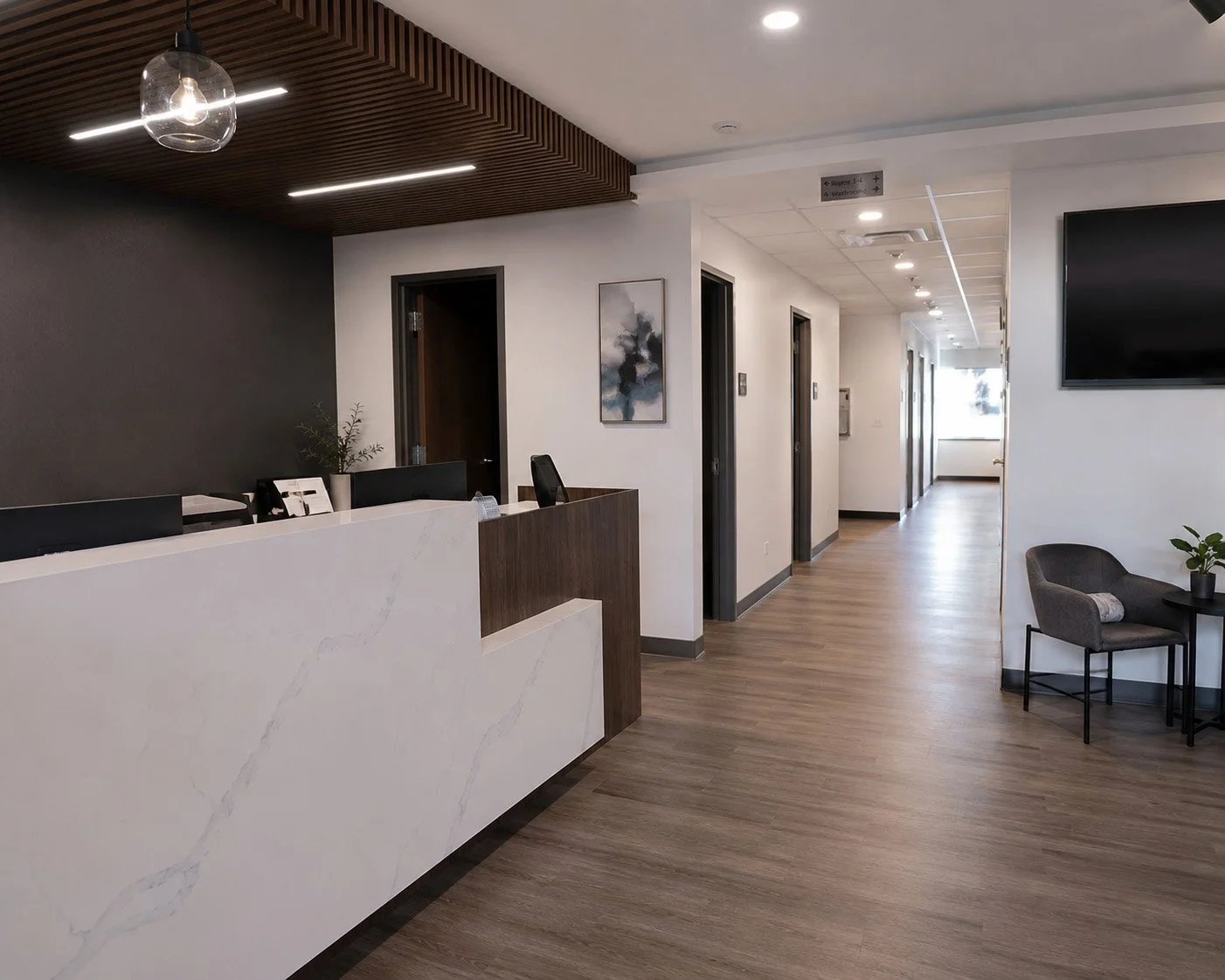 Reception area of a medical or office building with a white marble desk, a dark wall, and a hallway with several rooms. There is a chair with a pillow and a small table with a plant in the waiting area.